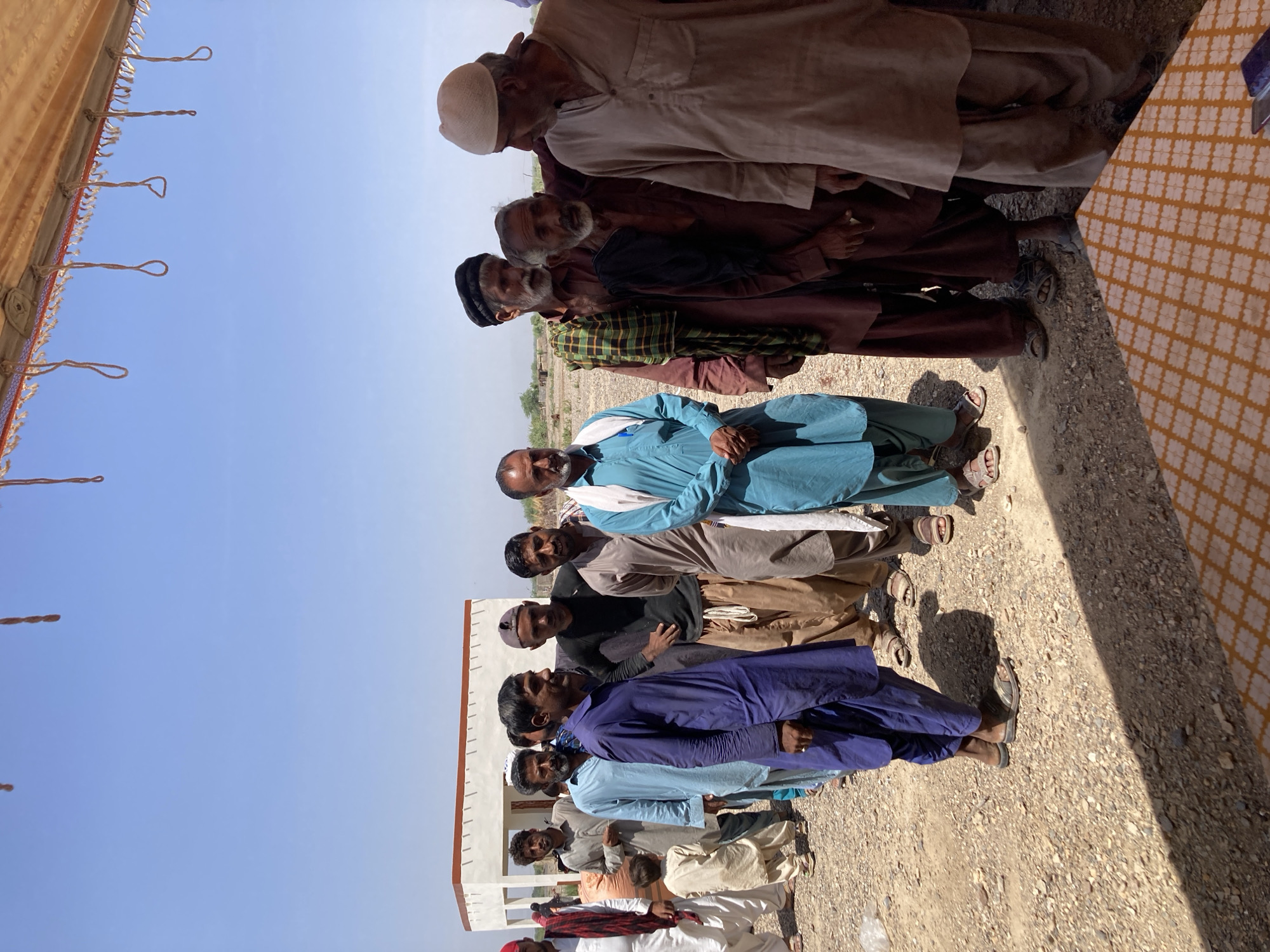 A group of men stands in a row in Hussenāni, Pakistan. The background consists of a cloudless blue sky and a gravel-covered ground. A building with a white facade and red trim is visible behind some of the men. A decorated fabric canopy hangs over them, providing shade. The men are dressed in a variety of traditional Pakistani clothing.