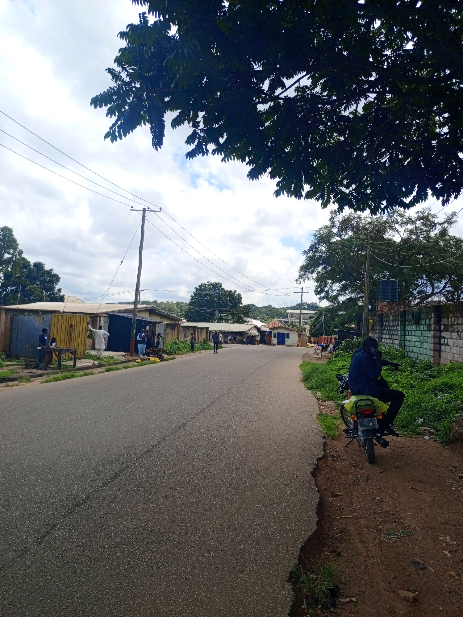 The image depicts a street scene in Jos, Nigeria, under a cloudy sky. The road, appearing to be asphalt, runs diagonally from the bottom left towards the middle distance, with a worn edge on the right side revealing a dirt embankment. On the left side of the road, several small buildings with corrugated metal walls are visible, some painted in yellow and blue. People are gathered in front of these structures, some appearing to be vendors or shoppers. One person in a white robe stands by a wooden table. On the right side of the road, a person is seated on a green motorcycle, wearing a dark jacket and pants. Behind the motorcycle, there is a brick wall, partially covered in graffiti, and lush green vegetation. Trees with dense foliage are present throughout the scene, with branches of a large tree extending from the top of the frame, casting shadows. Utility poles with overhead wires are also visible along the street. The overall impression is a busy, everyday street in a semi-urban area.