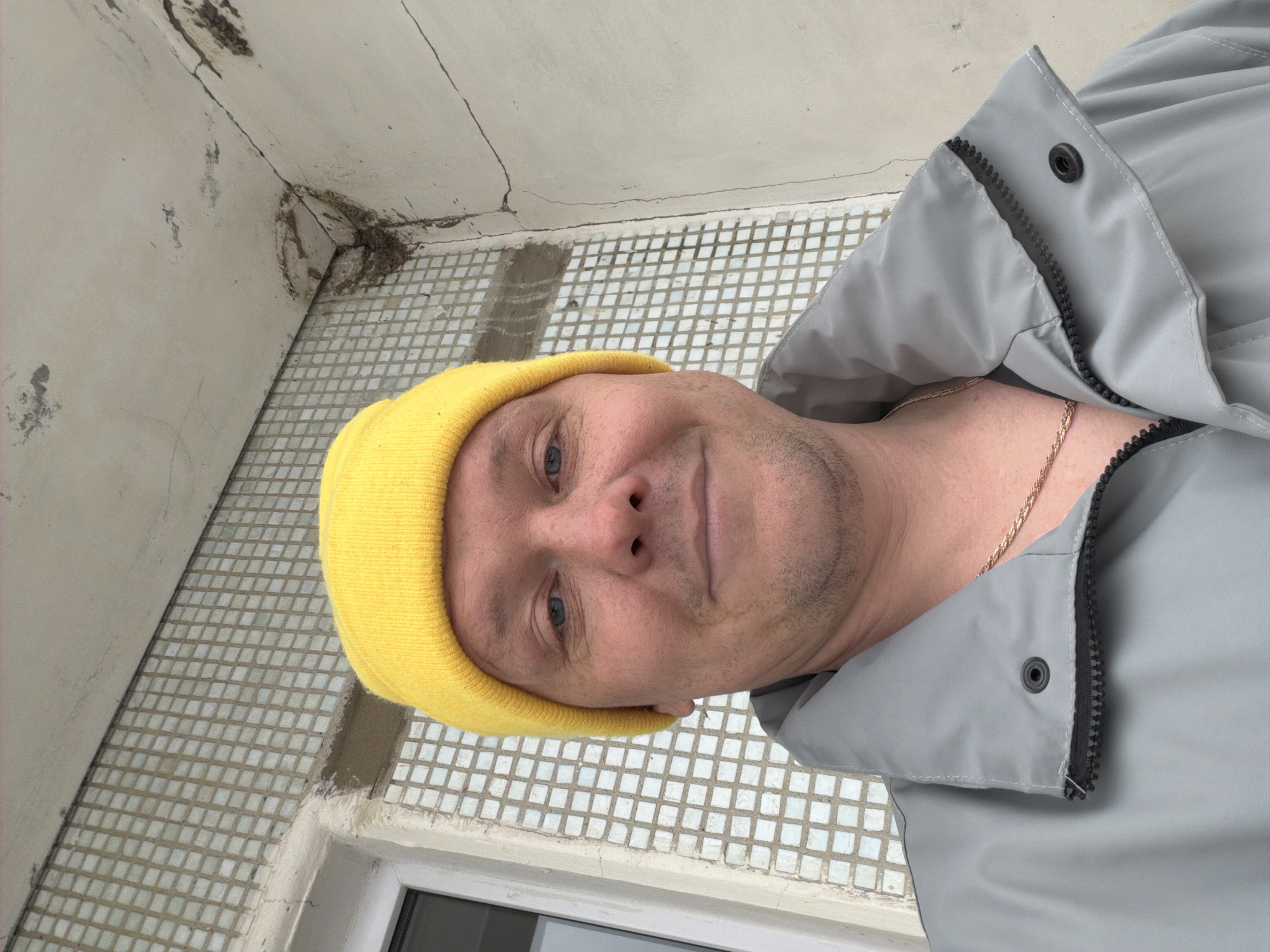 A close-up, high-angle photograph shows a man with fair skin, wearing a yellow beanie and a gray jacket with a black zipper, in a tiled corner of an unidentified location in Chaikovsky, Russia. The corner is tiled with small, light-colored tiles and has a window frame visible. A gold chain is visible around the man's neck. The wall is painted in a light color and shows signs of wear and tear.
