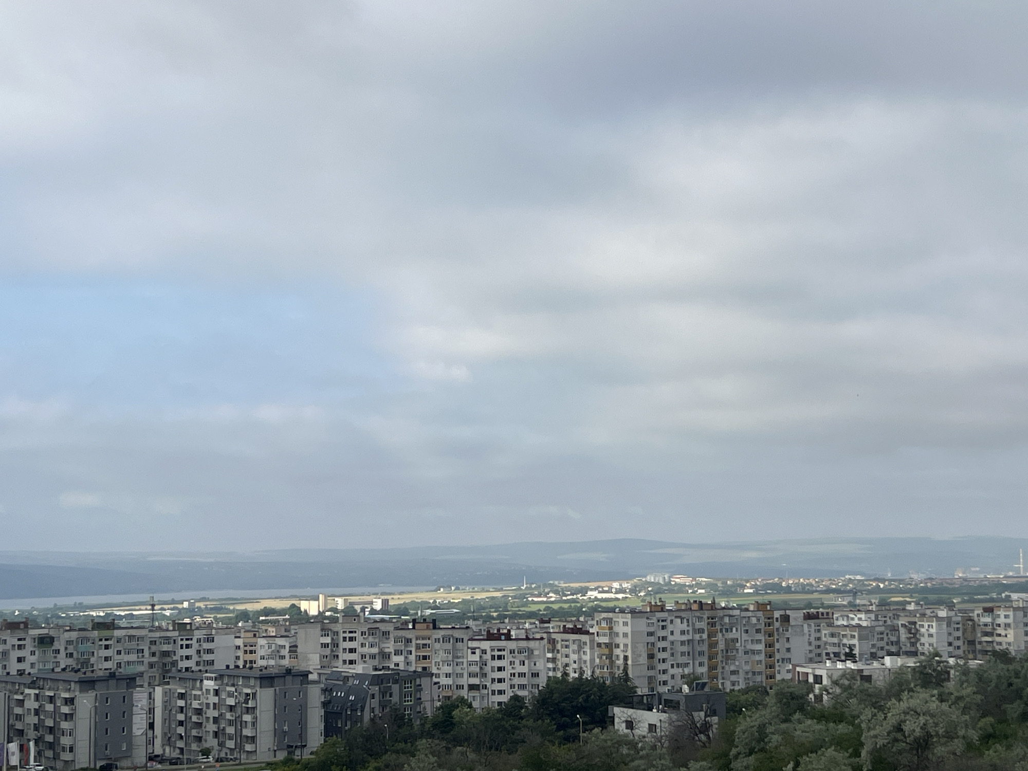 This is an eye-level, long shot photograph of a city skyline from a high vantage point in Varna, Bulgaria. The sky is overcast with grey and white clouds, with patches of light blue visible in the upper left quadrant. In the foreground, a dense cluster of apartment buildings dominates the lower half of the image. These buildings are mostly grey and white, varying in height, and appear to be residential structures. Some have visible balconies and window patterns.
Beyond the immediate apartment blocks, the city extends into the middle ground, with more buildings, some appearing shorter and lighter in color, interspersed with patches of green foliage and trees. In the distance, a body of water, likely the Black Sea, is visible as a hazy blue line, separating the city from a low-lying, undulating landscape of hills and plains that fade into the atmospheric haze. A thin, smokestack-like structure is visible on the right side of the distant horizon. The overall impression is of a sprawling urban environment under a diffused, cloudy sky. There are no people or discernible actions taking place in the scene.
