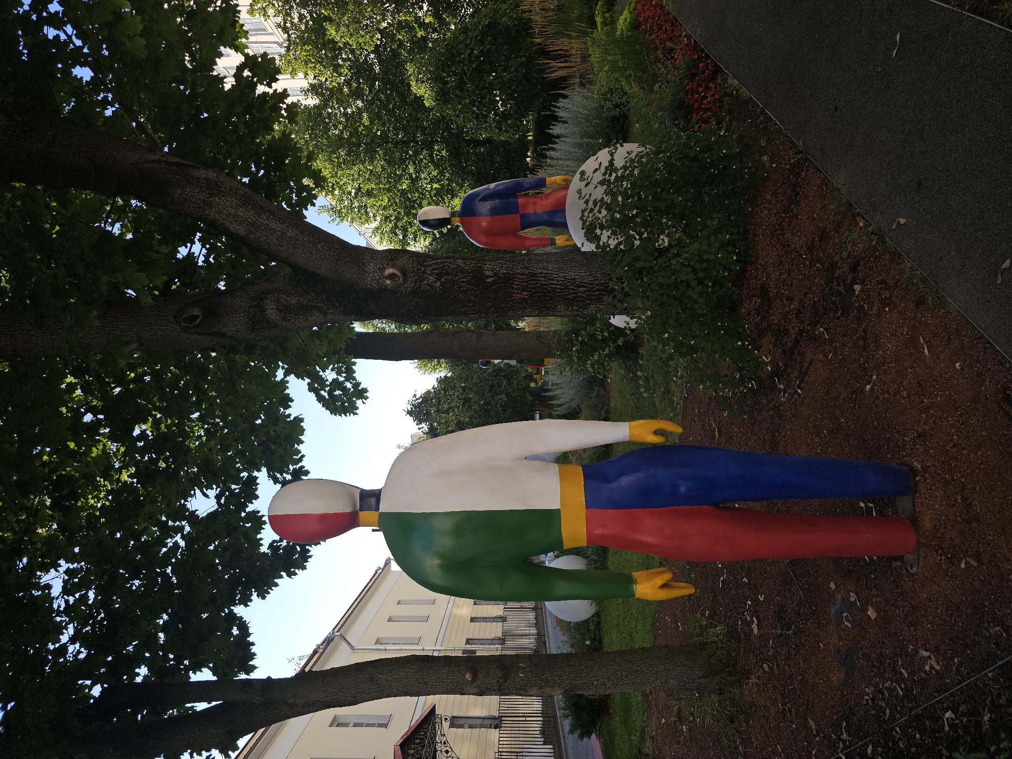 A full shot shows two colorful, life-sized abstract human sculptures standing in a park in Moscow, Russia, on a bright, sunny day. The sculpture in the foreground, facing away from the viewer, is predominantly white, green, and red, with yellow hands. It has a split design, with the left side of its body being green and white and the right side being red and white. Its legs are split into red and blue. The second sculpture, further back and to the right, is smaller and also abstract, with a similar split color design of red, blue, and white, and yellow hands. It is partially obscured by foliage and a large white rounded object.
The sculptures are situated in a park with lush green trees, some with thick, textured bark. The ground is covered in dark brown mulch. In the background, there is a light-colored building with multiple windows and a black wrought-iron fence. The sky is clear and blue. A paved walkway runs diagonally across the lower right of the frame, bordered by a flowerbed with red and dark green foliage. The overall impression is one of an artistic installation in an urban green space.