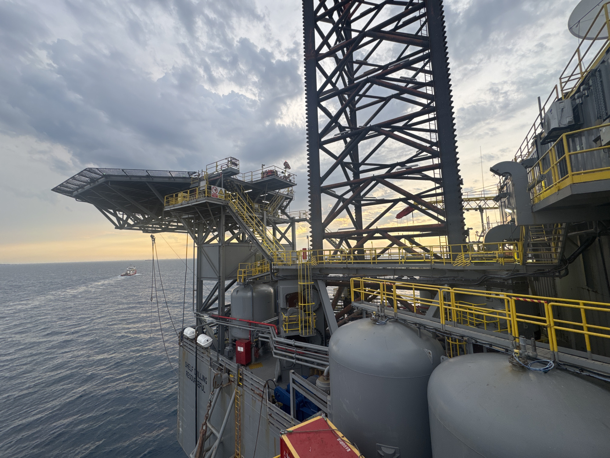This is an outdoor, eye-level, medium shot of an offshore drilling rig and the surrounding ocean under a cloudy sky. The sun is low on the horizon, casting a warm glow. The rig is a complex structure of metal beams, platforms, and equipment, including a helideck. Two large, grey storage tanks are visible on the lower deck. A small boat is moving away from the rig in the distance. The water is a deep blue with moderate waves. The image is static, and there are no people or animals visible. The location is an offshore drilling rig, not in a city.