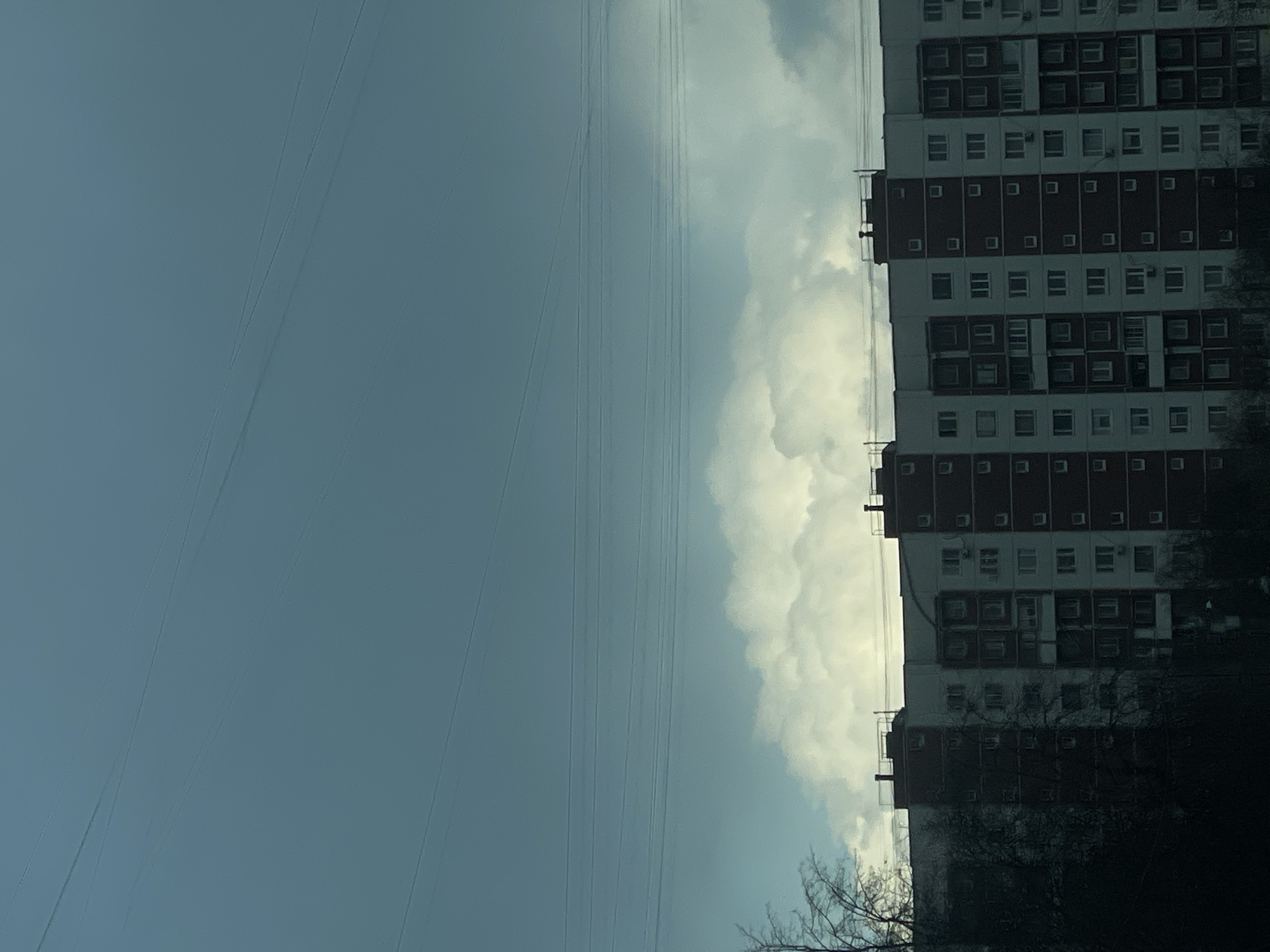 The image shows a tall building next to a partly cloudy sky with overhead power lines. The building appears to be an apartment complex, with numerous windows on each floor. The power lines run horizontally across the sky, between the building and the clouds. Trees and vegetation are visible near the base of the building. The exact location within the city of Unknown, Unknown, cannot be determined from the image.