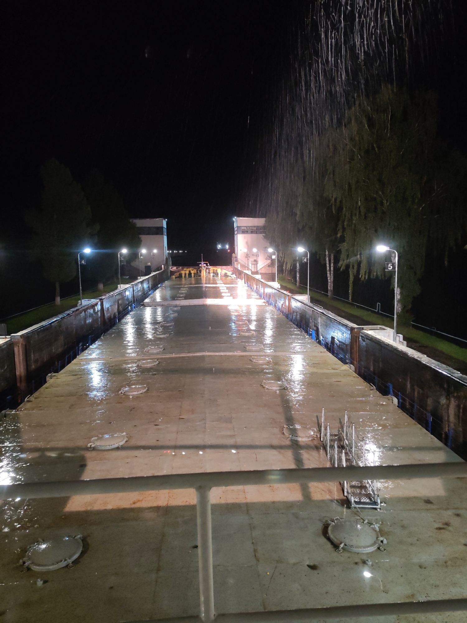 This is a nighttime, eye-level, slightly elevated, full shot of a rain-slicked canal lock with a barge or ship deck in the foreground. The scene is illuminated by artificial lights, including streetlights and lights from the lock structures, which cast reflections on the wet surfaces. The rain is visible as streaks falling from the sky.
In the foreground, the deck of a vessel is visible, with several circular hatches, some partially submerged in water. A white metal railing runs along the edge of the deck. In the middle ground, the canal lock walls are visible, lined with what appear to be blue ropes. The lock itself is filled with water, reflecting the lights. A metal walkway or gangway with railings is positioned on the deck.
In the background, the lock gates and associated structures are illuminated. Trees line the sides of the canal, their leaves glistening in the rain. Streetlights with numbers "4" and "5" are visible along the canal edge. The overall impression is a functional, industrial setting experienced during inclement weather. The location is identified as a canal lock in Markovo, Russia.