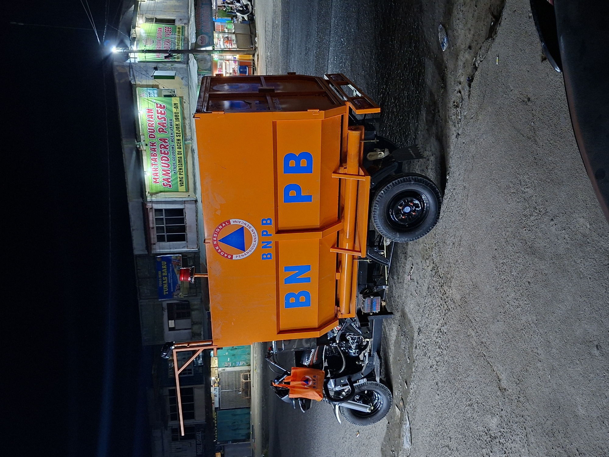The image shows an orange vehicle with the acronym "BNPB" painted on its side in blue, an acronym commonly used by the Indonesian National Board for Disaster Management, parked on a paved street in Keudegeudong, Indonesia. The vehicle appears to be a modified motorcycle with a cargo box attached. To the left of the vehicle, several buildings are visible, including a storefront with the signage "MARTABAK DURIAN SAMUDERA PASEE," which advertises durian martabak, a local dessert. The image was taken at night, with artificial lighting illuminating the street and buildings. A portion of another vehicle is seen to the right of the frame. The overall impression is of a street scene with a disaster management vehicle parked in front of local businesses at night.