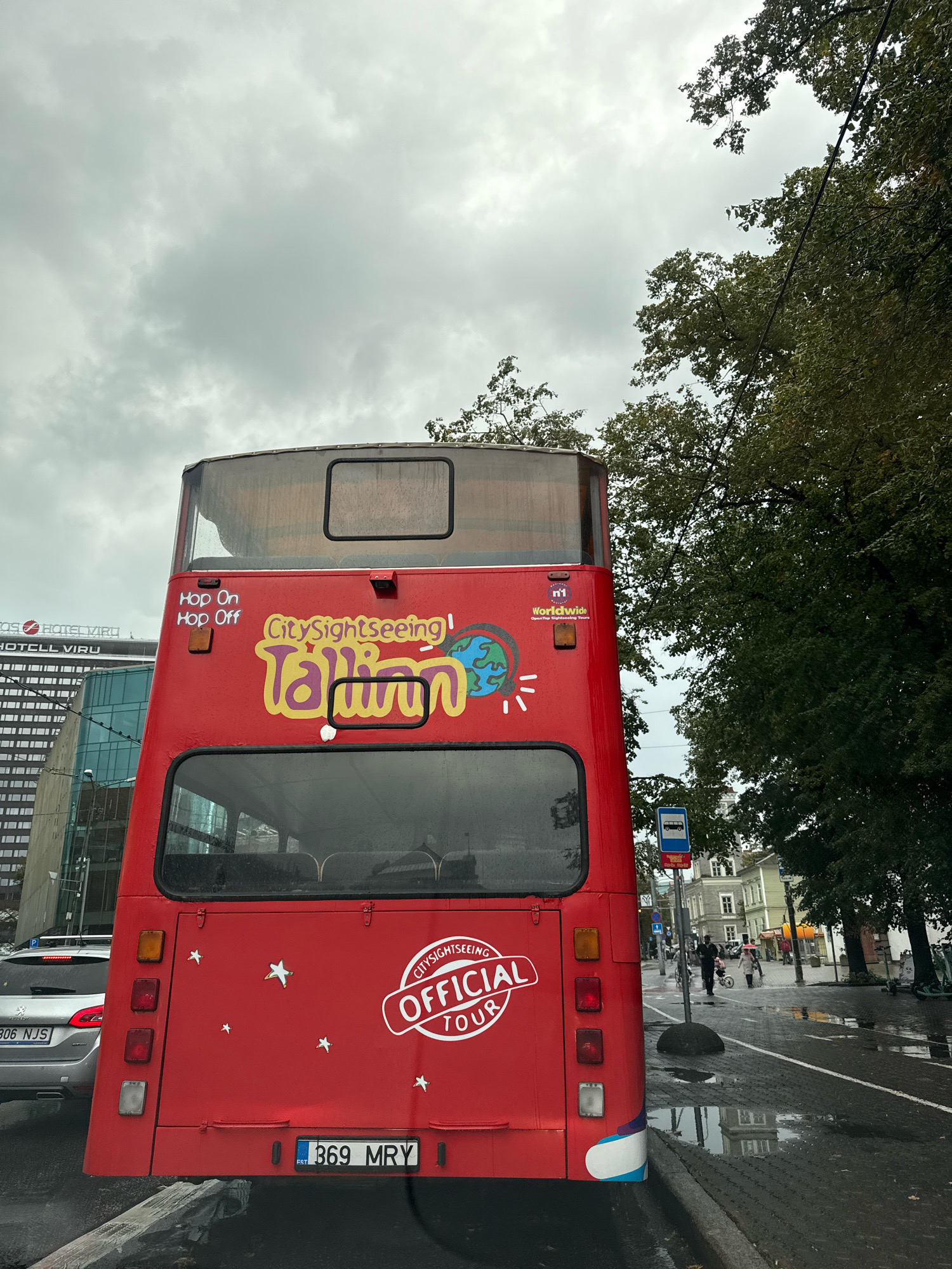 This is a rear view of a bright red double-decker bus with "CitySightseeing Tallinn Official Tour" written on its back in colorful lettering. The bus has a "Hop On Hop Off" sticker on its upper rear left side. It has multiple tail lights and a license plate with "369 MRY" and "EST" visible. The bus is parked on a wet city street, possibly in Tallinn, Estonia, as indicated by the bus's branding. In the background, there are trees, buildings, a silver car with a license plate ending in "NJS", a bus stop sign, and several people walking on a sidewalk. The sky is overcast and cloudy, and there are reflections in puddles on the street, suggesting it has recently rained or is raining.