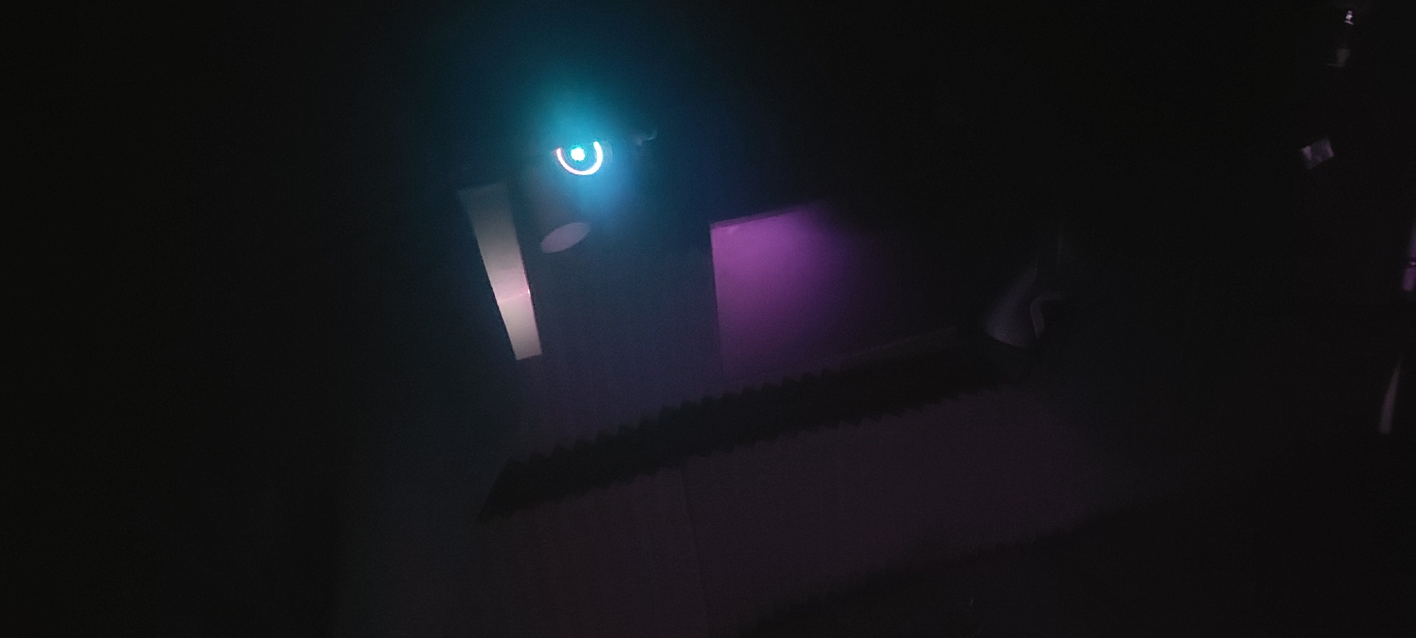 The image depicts a dimly lit interior, possibly a room. There is a circular light source emitting a blueish-green glow on the upper left, suggesting an electronic device or a small lamp. Adjacent to this, a rectangular surface reflects a soft, white light. Another rectangular surface in the center emits a faint, purple glow. A surface with a serrated edge is visible below these light sources, perhaps a piece of equipment or a design element. The rest of the scene is shrouded in darkness, making it difficult to discern further details. The image's composition and lighting suggest a modern, potentially technological environment. The precise location is unknown, but it is said to be in Tashkent, Uzbekistan.