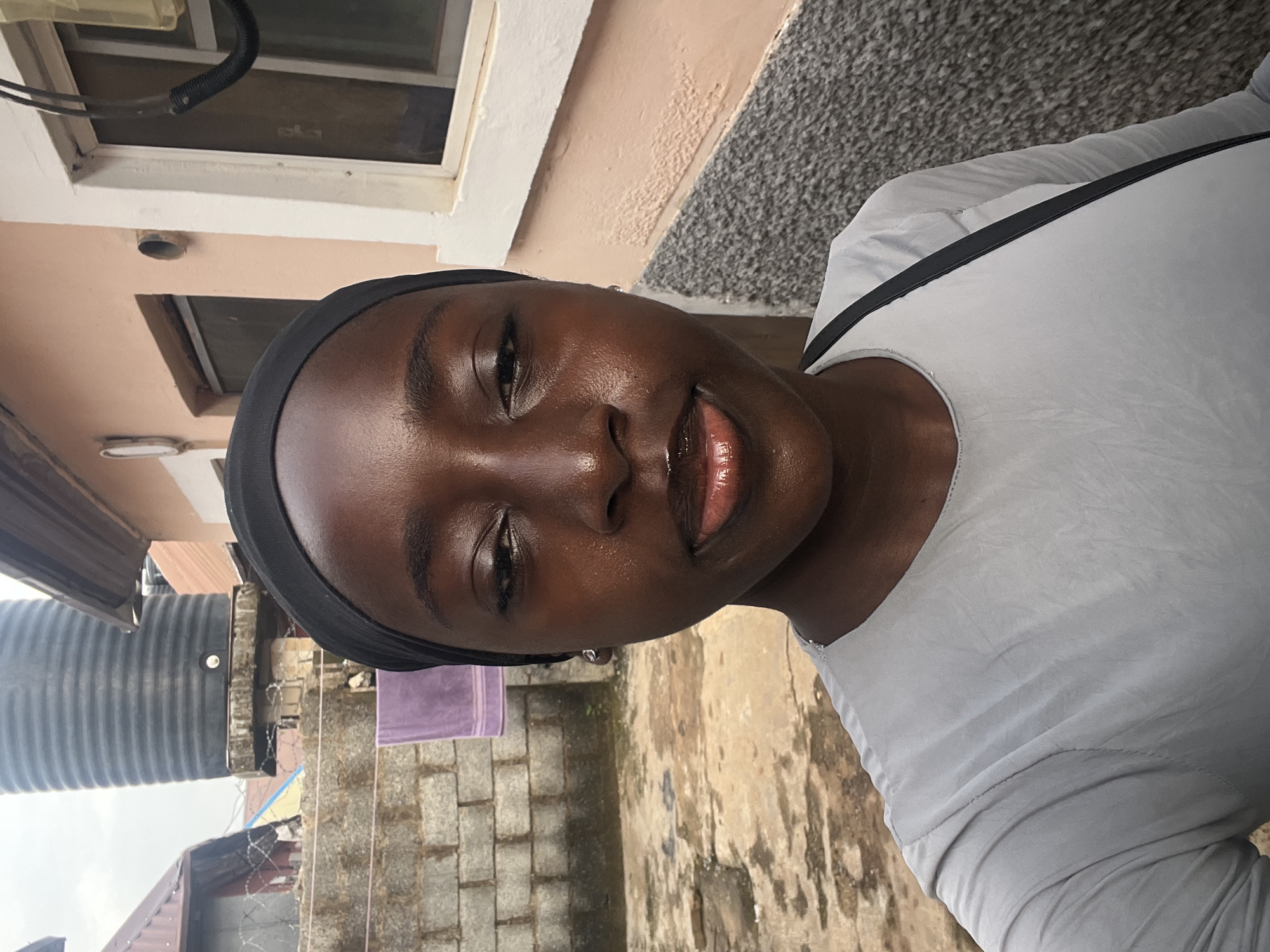 A young Black woman wearing a grey shirt and a black headscarf is shown in a selfie. She has dark skin, and her lips are shiny with lip gloss. In the background, there is a brick wall, a large black water tank, and a building with a peach-colored exterior and a window. A purple towel is hanging on the brick wall. The scene appears to be an outdoor setting, possibly a residential area in Abuja, Nigeria.