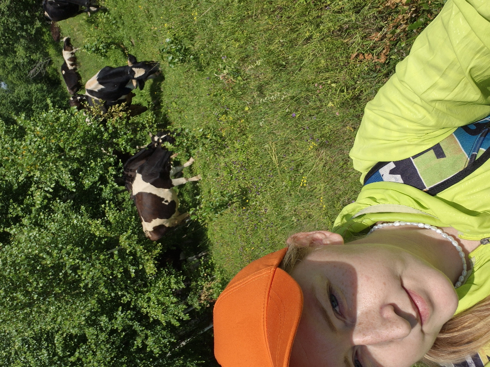This is a vertical, high-angle, outdoor photograph taken on a sunny day in Karsha, Russia. The main subjects are a young person and several cows. The person, partially visible in the foreground, is lying on their back and wearing an orange baseball cap and a bright lime green jacket with a colorful strap across it. They have fair skin with freckles and are looking towards the camera with a slight smile. A pearl necklace is visible around their neck.
In the background, several black and white Holstein cows are grazing on a grassy field dotted with small yellow and purple wildflowers. Some of the cows are partially obscured by dense green bushes. The overall scene suggests a rural, pastoral setting. The lighting indicates it is daytime, likely mid-morning or afternoon, given the strong shadows. No visible text is present in the image.