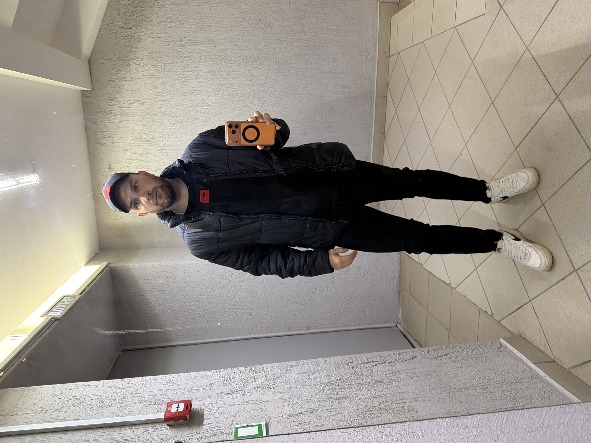 A man stands in a hallway, taking a selfie with an orange smartphone. He is wearing a black puffer jacket, black pants, and white sneakers. He has a dark beard and is wearing a baseball cap with a red brim. The hallway has tiled floors and textured walls. A fire alarm and a sign are visible on the wall. The location is implied to be in the city of Chaikovsky, Russia, though this is not explicitly confirmed by any visual element in the image.