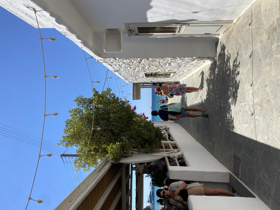 The image depicts a sunlit, narrow, stone-paved alleyway leading towards a coastline in a Greek town, identified as Φάρος. The alley is flanked by whitewashed buildings. On the left, an outdoor dining area under a wooden canopy, with visible patrons and a low white wall, abuts a large green tree with pink floral accents. On the right, a textured white stone building features a recessed opening and a closed, light-colored wooden door. Overhead, multiple strings of bare bulb lights and utility lines are visible against a clear blue sky. In the foreground left, a female subject in sunglasses and light-colored clothing is observed interacting with a mobile phone. In the mid-distance, two subjects, a male and a female, are walking away from the viewer towards the ocean. The male wears a dark shirt, patterned shorts, and carries a light blue tote bag. The female wears a colorful patterned dress and a red hat. The background reveals a turquoise sea, a sandy beach with indistinct figures, and several large white yachts. Shadows are cast upon the pavement from the structures and tree.