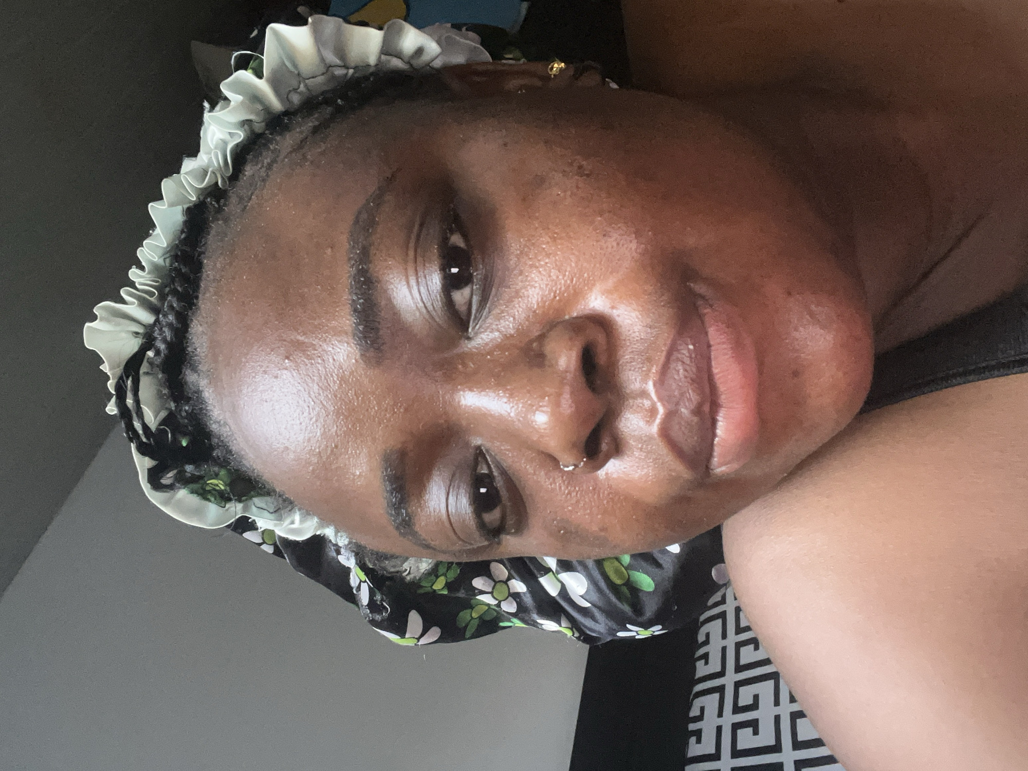 A medium shot of a dark-skinned individual's face with a nose ring and a white ruffled bonnet. The individual is lying down with the head turned sideways. The background is out of focus with a black and white patterned fabric. The location cannot be determined.