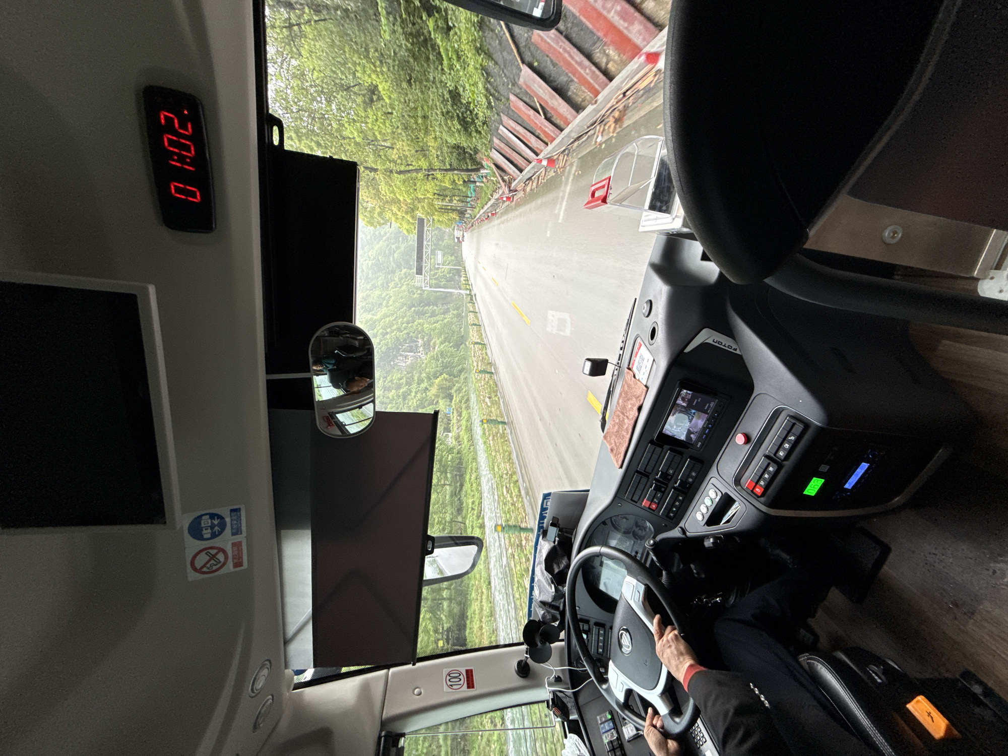 The image shows the view from the driver's seat of a vehicle, likely a bus, traveling on a road surrounded by green foliage. The time displayed on a digital clock in the vehicle is 01:02. The driver's hands are visible on the steering wheel. The location is likely in the Wulingyuan area of China, based on the type of roads and vegetation.