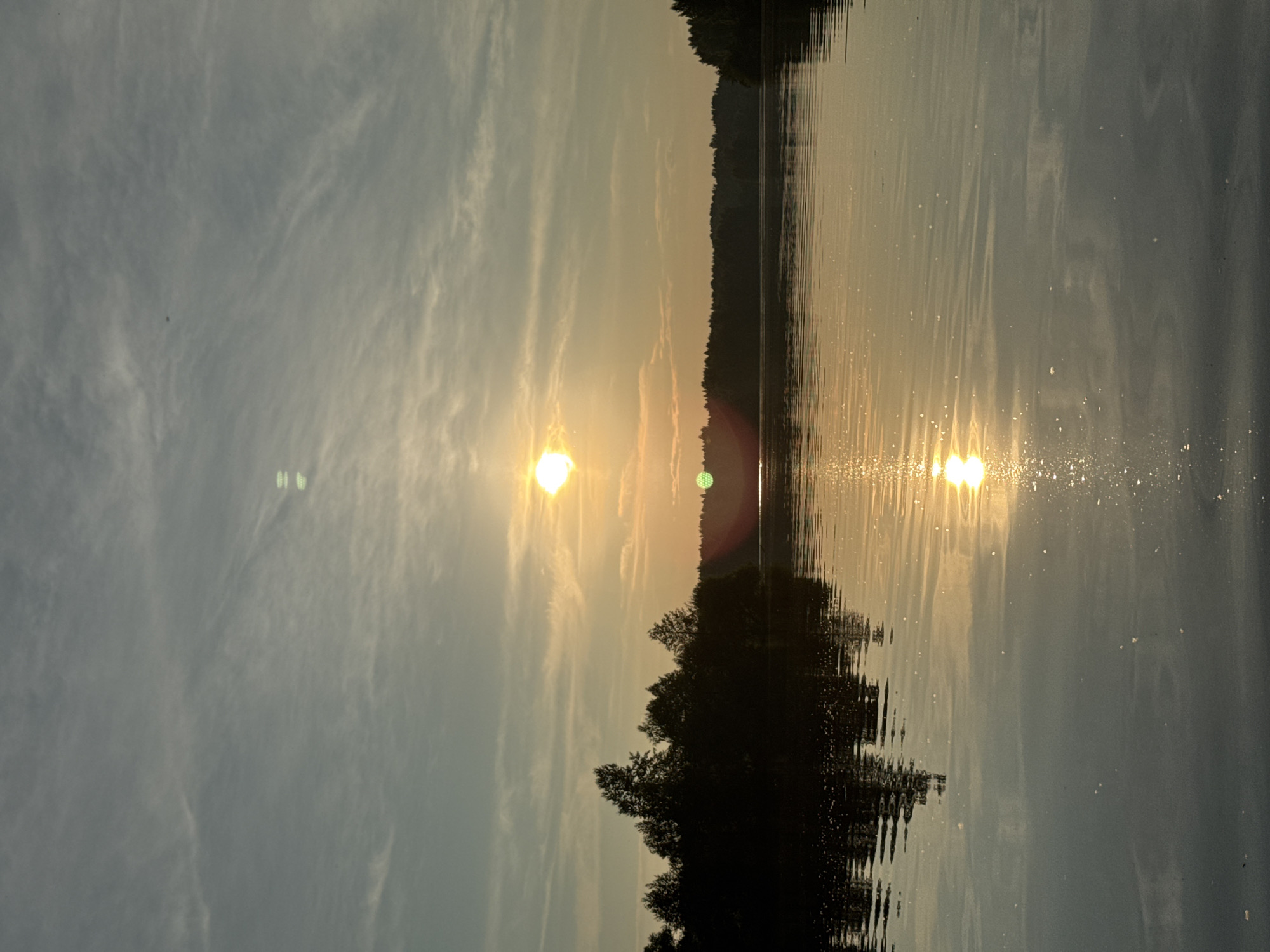 The image depicts a serene sunset over a body of water, likely a river or lake, with a silhouetted treeline in the background. The sun, a bright orb of golden light, is positioned slightly left of the center of the frame, casting a warm glow across the sky and reflecting on the water's surface. Wispy clouds are scattered across the pale blue and orange sky. The water is calm, with subtle ripples that catch the sunlight, creating shimmering patterns. The dark silhouette of trees lines the far bank, creating a stark contrast with the illuminated sky and water. The image appears to be taken from a watercraft, as the perspective is low and close to the water. The scene evokes a sense of tranquility and natural beauty. The location in Kamysh, Russia, is not explicitly identifiable within the visual information provided.