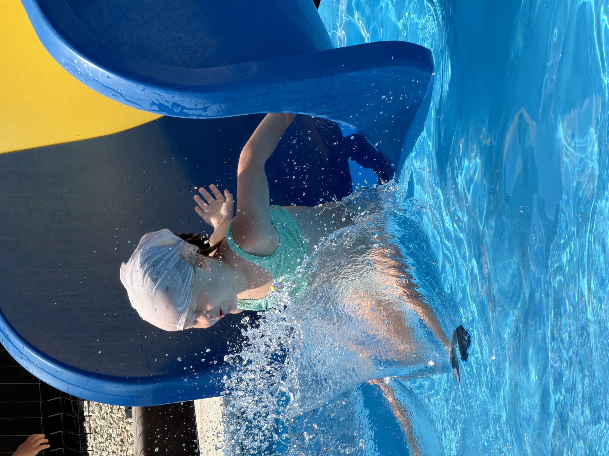 A young girl, wearing a teal swimsuit and a white swim cap, slides down a blue water slide and splashes into a bright blue swimming pool. The sun is shining, casting dappled reflections on the water's surface, suggesting a bright, warm day. She is captured mid-splash, with water droplets flying around her. Her eyes are closed, and her expression seems to convey excitement or perhaps surprise at the impact. The water slide has a yellow section at the top. In the background, a black fence and some gravel are visible, hinting at an outdoor recreational area, possibly a water park or a backyard pool. The scene is set in Kamyshin, Russia. There is no visible text in the image.