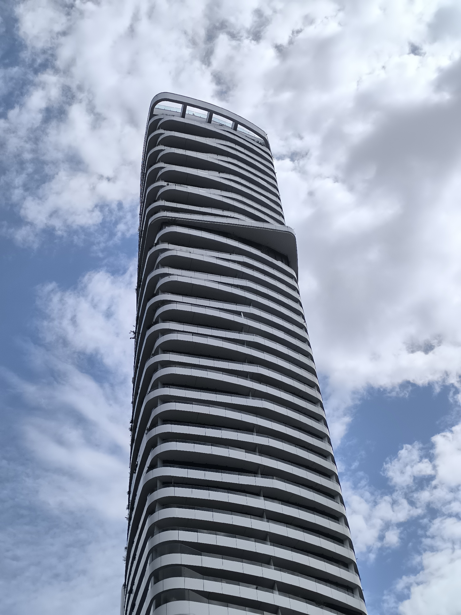 The image shows a modern, twisting skyscraper against a cloudy blue sky. The building features a white, undulating facade with numerous balconies that appear to wrap around the structure. The balconies are delineated by horizontal white bands. Towards the top, the building culminates in a more angular, open-air observation deck or terrace. The perspective is from a low angle, looking up at the towering building. The sky is filled with a mix of white and grey clouds, suggesting a dynamic weather day. There are no visible people or vehicles in the frame. The building appears to be residential or commercial, with a sleek, contemporary architectural style. The context of Vienna, Austria, is not directly evident from the image itself, but based on external information, this building is the 'TrIIIple' residential tower in Vienna.