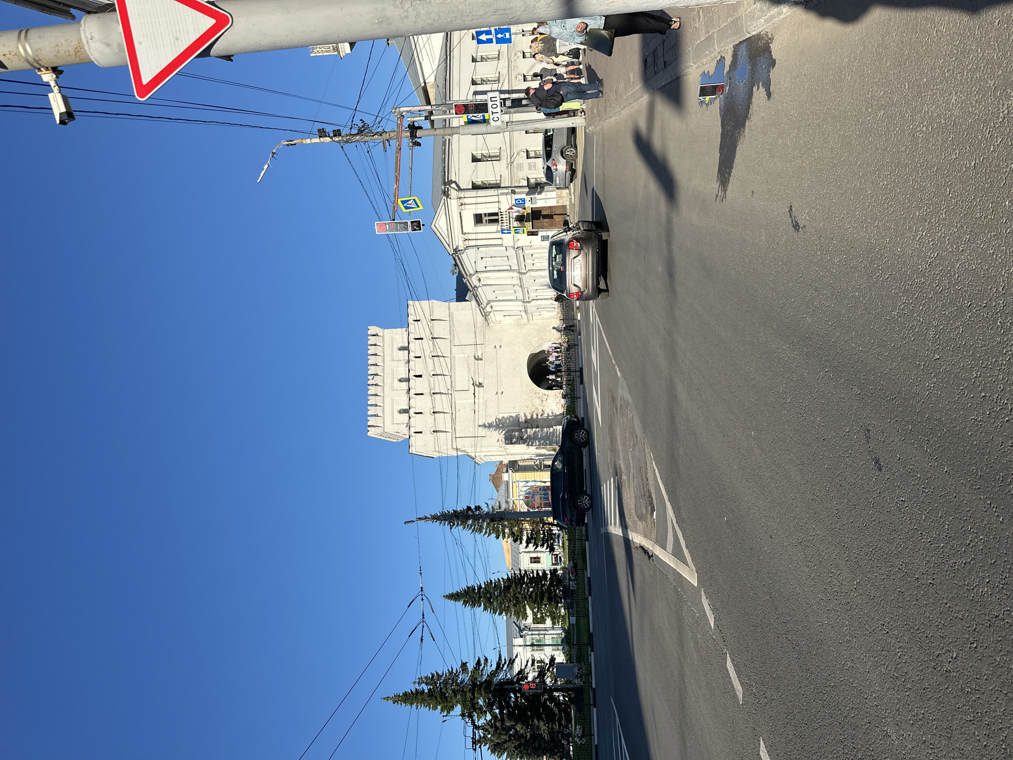 The image captures a view of a street in the city of Feklino, Russia, on a clear, sunny day. In the background, a large, white, crenellated structure, likely a historical gate or tower, dominates the scene. In front of this structure, a group of people are visible near an arched entryway. To the right of the tower, a white building with multiple windows is situated.
The foreground of the image shows a paved road with white lane markings. Several cars are present on the road; a dark SUV is parked on the left side, and a silver sedan is driving away from the viewer in the center-right. A crosswalk is marked on the road.
Along the right side of the road, there is a sidewalk with several pedestrians. A tall, gray utility pole with various wires, a security camera, and traffic signals is prominently featured. A red and white triangular road sign, indicating a yield or give way, is attached to this pole. Other road signs, including a blue directional sign with an arrow and a white sign with the word "СТОП" (STOP in Russian), are also visible.
To the left of the road, there are several tall evergreen trees. Traffic lights are also visible, with one showing red. The sky is a vibrant, clear blue. The overall impression is of an urban environment with historical architecture and active street life.