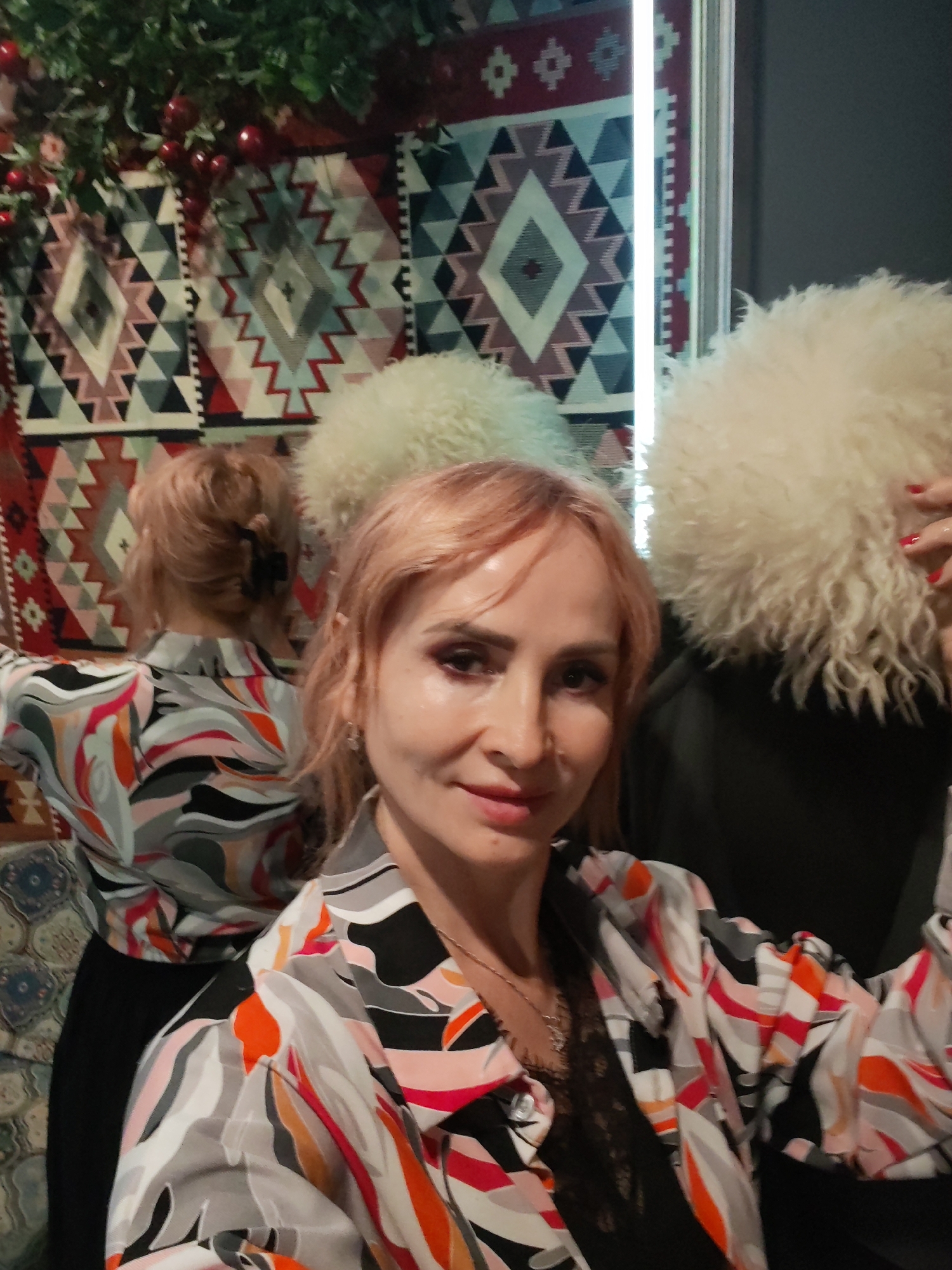 The image shows a woman with light pink hair taking a selfie. She is wearing a black lace top underneath a colorful button-down shirt with a swirling, abstract pattern in shades of grey, black, orange, pink, and white. She has on dark eye makeup and her lips are painted a muted red. Her hair is styled in loose waves, with some pulled back from her face.
In the background, there is a patterned tapestry or rug with geometric designs in red, black, white, and grey, possibly with a diamond motif. To the right of the woman, there is a large, fluffy white hat that appears to be made of fur. Another similar hat is partially visible behind her to the left. A reflection in a mirror or glass surface shows the back of the woman's head and shoulders, also wearing the patterned shirt, and a hint of the tapestry and the white hat. Green foliage with red berries is visible in the upper left corner, suggesting a decorative element or perhaps a seasonal display.
The lighting suggests an indoor setting, possibly a restaurant or a decorated space. The mood appears casual and relaxed. There is no visible text in the image. The context of Yoshkar-Ola, Russia, does not provide specific visual cues within the image itself.