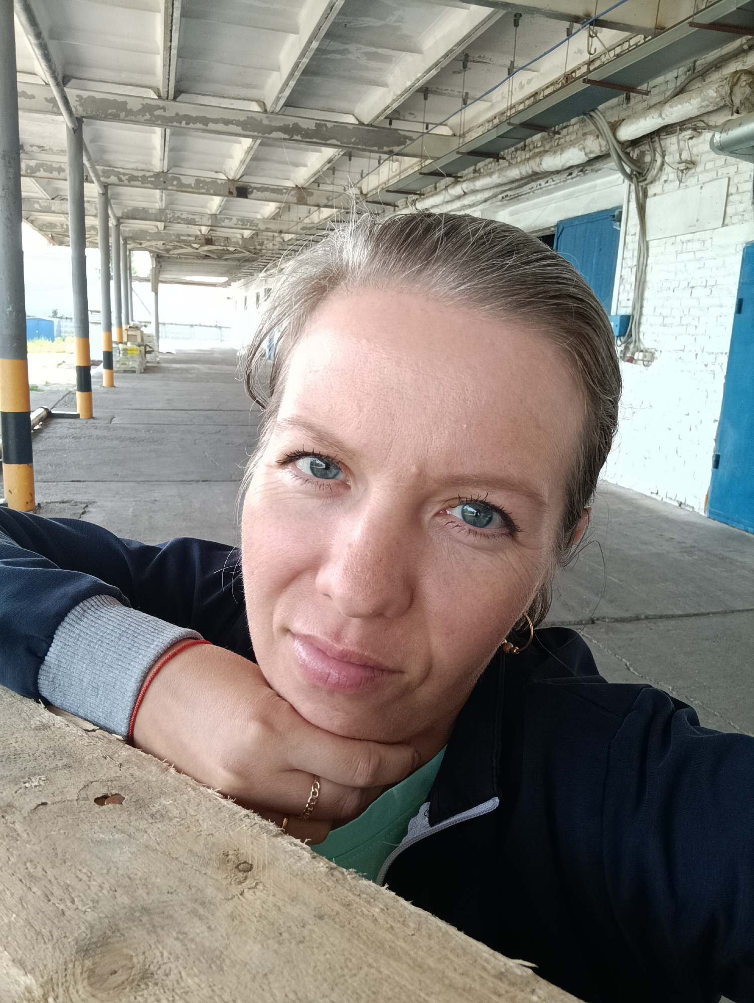 A woman with light skin, graying blonde hair pulled back, and blue eyes is the main subject of this selfie. She is wearing a dark blue jacket with a gray ribbed cuff and a thin red bracelet on her wrist. A gold chain ring is visible on her finger. She is resting her chin on her hand, which is resting on a rough, light-colored wooden surface.
The background suggests an outdoor, industrial-type location. A covered walkway is visible, supported by gray pillars marked with black and yellow stripes. The walkway's ceiling appears to be concrete or a similar material with visible seams. In the distance, the walkway leads towards a brighter area. To the right of the woman, a white brick wall is visible with two closed blue doors. Wires and pipes are visible along the top of the wall.
The location is the city of Chaikovsky, Russia.