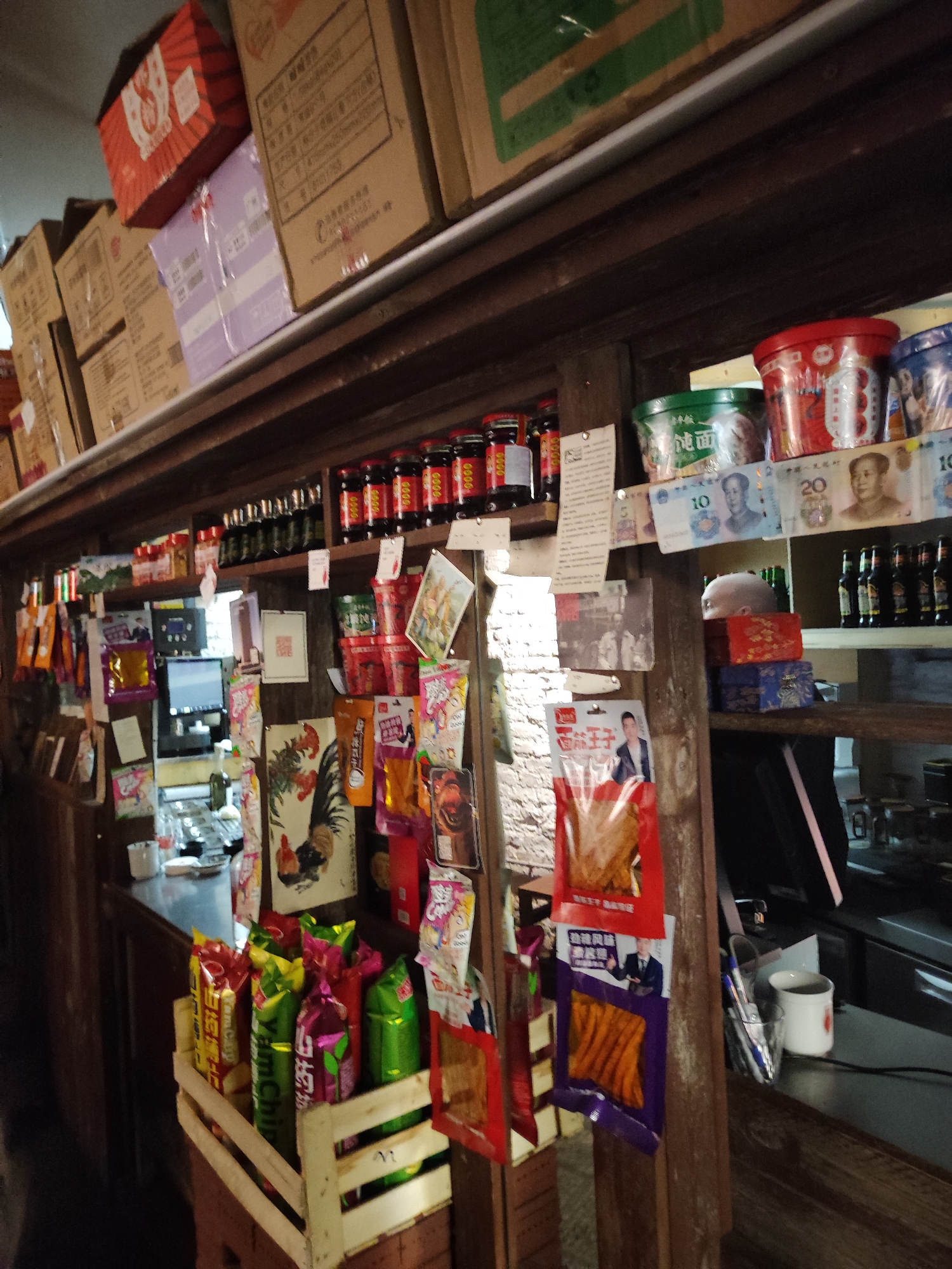 This indoor photograph captures a cluttered and rustic-looking store interior, likely a convenience shop or a small grocery. The scene is dominated by wooden shelving, filled with various packaged food items, bottles, and jars, suggesting a compact retail space.
In the upper section, large cardboard boxes are stacked on shelves. Below, numerous small glass bottles with red caps are neatly arranged. The middle and lower shelves display a wide assortment of snacks, instant noodles, and other goods. Several cups of instant noodles, some red and some green, are visible. Chinese Yuan banknotes, specifically 10 and 20 denominations featuring Mao Zedong's portrait, are creatively displayed as decoration near the instant noodles. A counter is visible on the right, with a computer monitor partially obscured and a white mug resting on it. A wooden crate filled with brightly colored snack packages, including green, pink, and red bags, sits on the floor or a lower platform. Decorative items, such as a paper cut-out of a rooster and various small posters or notes, are affixed to the wooden supports.
There is no discernible human activity or event taking place; the image presents a static view of the store's arrangement. The lighting appears somewhat dim but adequate, creating a cozy and perhaps slightly aged atmosphere. The numerous Chinese characters on product packaging, coupled with the Chinese currency, strongly indicate a location within China or a Chinese-speaking region.
Visible text includes: "0000" on bottle caps, "Yam Cary," "Yam Chip," "纯面" (pure noodles), "面筋王子" (Gluten Prince), and "C" on a box. The banknotes clearly show "10" and "20."