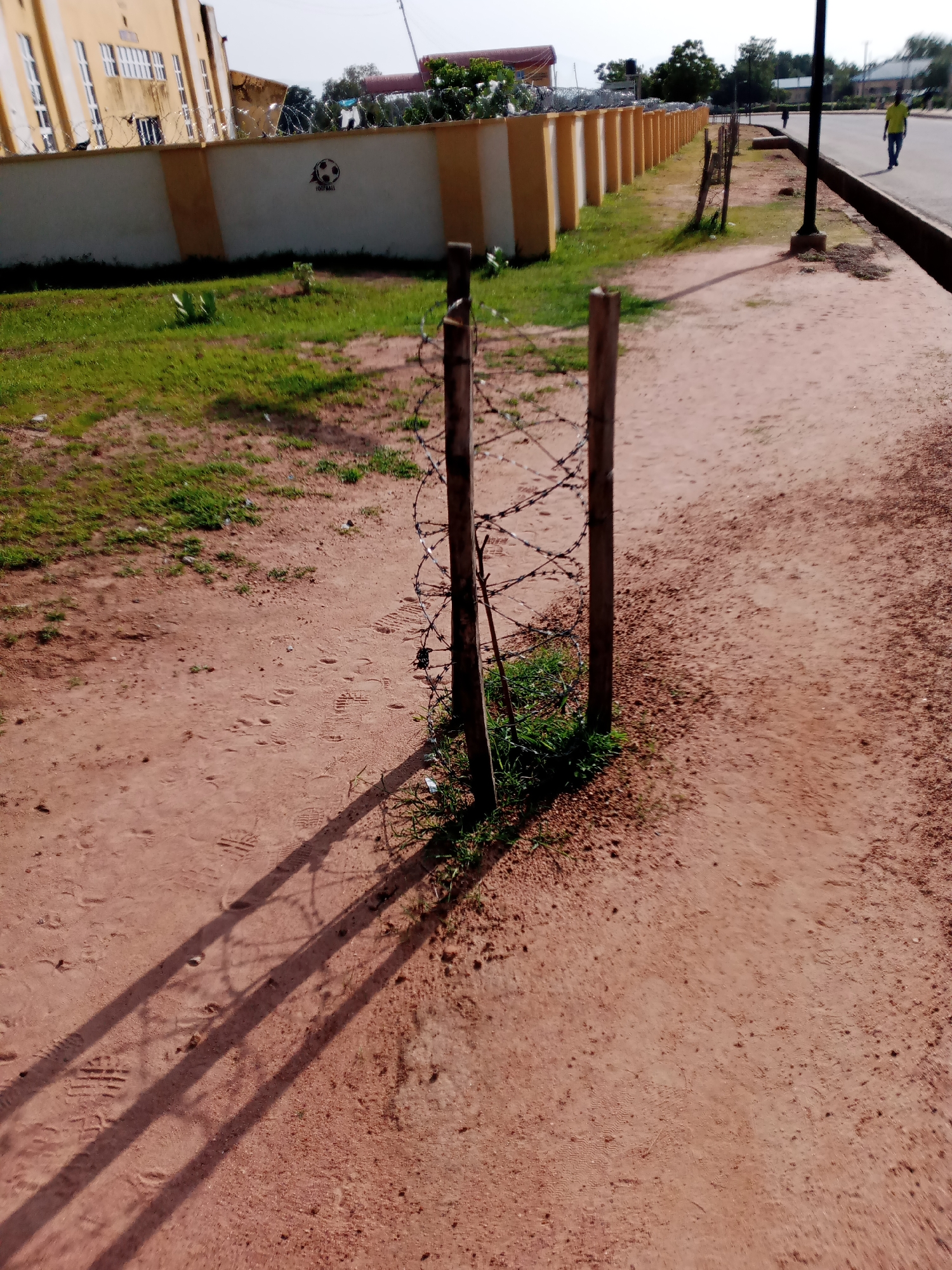 A street scene in Jalingo, Nigeria, features a sandy path bordering a grassy area with a low wall topped with barbed wire. Two wooden posts supporting the barbed wire fence are prominent in the foreground, casting long shadows on the dirt path. Behind the wall, a building with yellow and white sections and barred windows is visible. In the background, a street with a sidewalk can be seen, with two individuals walking on it, one in a yellow shirt and the other in a dark shirt. The sky is bright and clear.