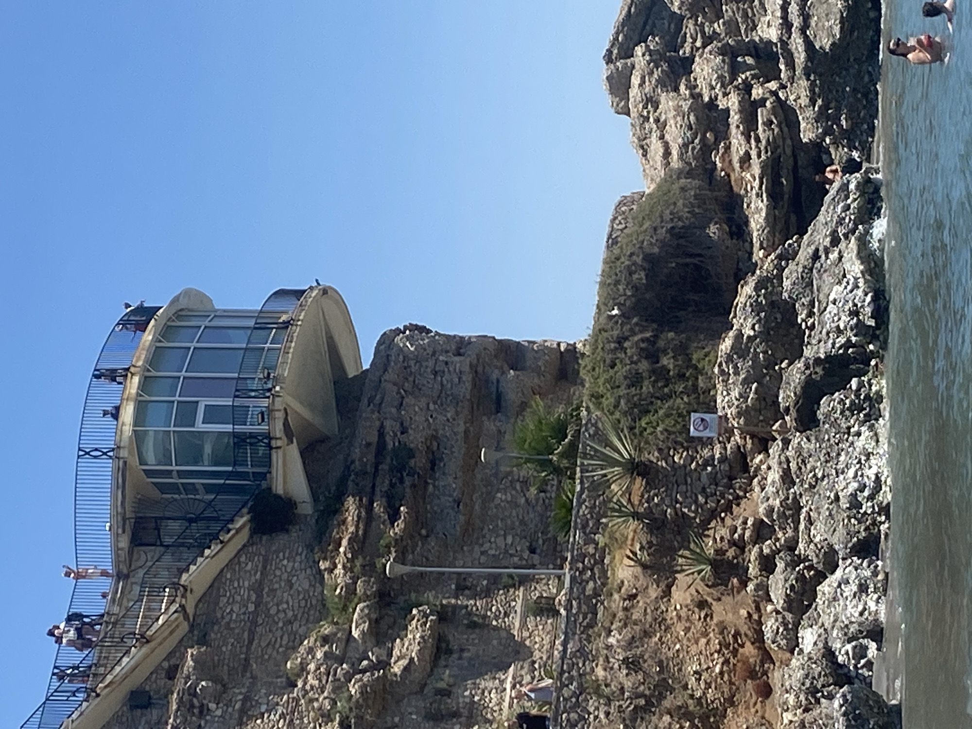 The image shows a view of the Balcón de Europa in Nerja, Spain, on a sunny day. A modern glass-enclosed structure with a rounded balcony juts out from a rocky cliff face. A curved staircase with black metal railings leads up to the balcony, where several people are visible standing and looking out. The cliff itself is rugged, with stone retaining walls and sparse vegetation, including agave plants. Below the cliff, the dark water of the sea laps against a rocky shore. In the water, several people are seen swimming and relaxing, with one woman in the foreground holding a drink. The sky is a clear, bright blue.