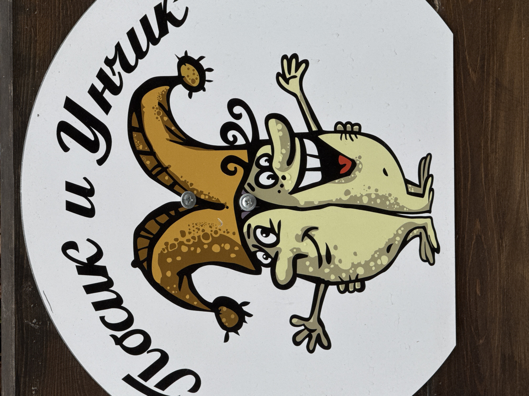 The image shows a round sign affixed to a wooden surface, possibly a wall or door. The sign is white with a cartoon illustration of two conjoined, comical characters. One character wears a jester's hat and has a mischievous expression. The other character has a wide, toothy grin and is waving. The sign also features Cyrillic text, which translates to "The Sly and Clever." The style of the illustration is reminiscent of children's book art. The image was taken in the city of Perm, Russia.