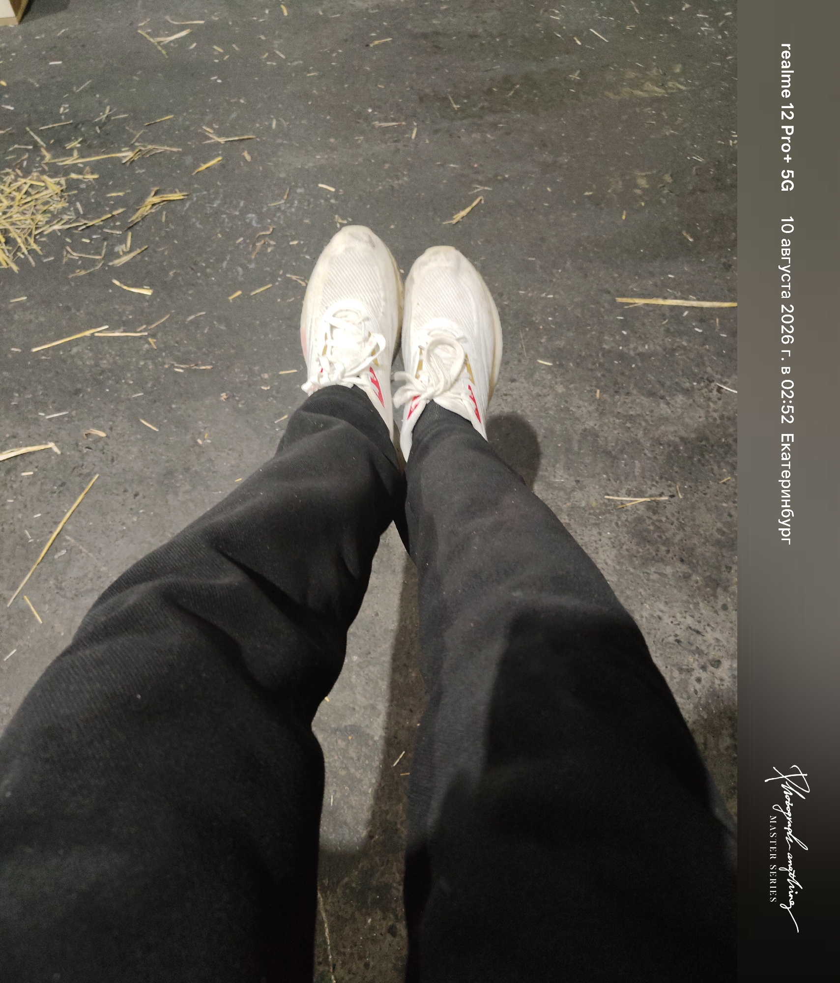 The image shows a person's legs and feet from a first-person perspective, wearing white athletic shoes with red accents and black pants. The setting appears to be an outdoor ground surface with scattered straw or hay. A timestamp on the right side indicates the photo was taken on August 10, 2026, at 02:52 in Yekaterinburg, Russia. The text "realme 12 Pro+ 5G" and a stylized signature with "MASTER SERIES" are also present.
