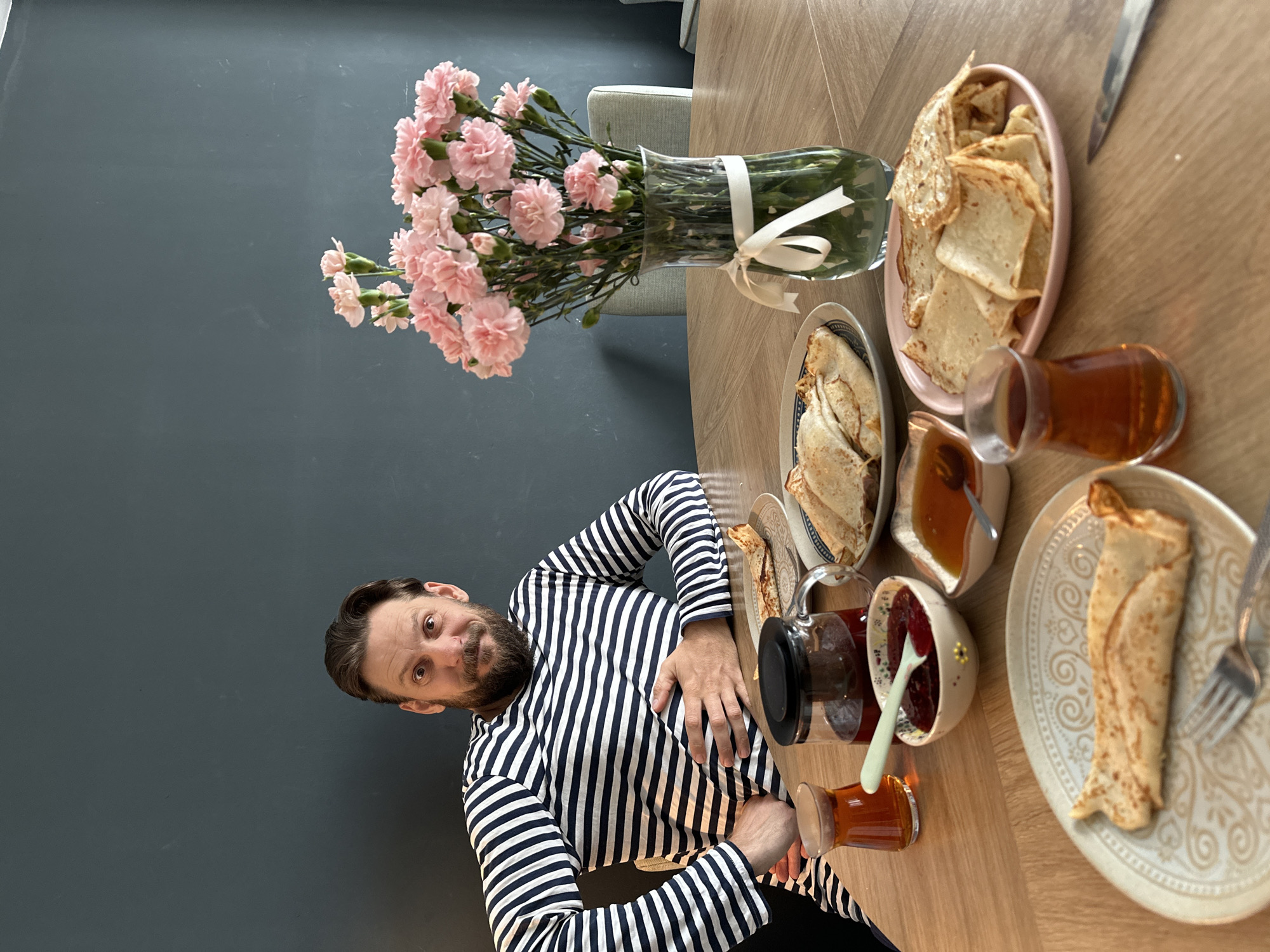 A photograph taken indoors shows a man seated at a table laden with food and drinks. The man, sporting a striped shirt, is positioned to the left of the frame. He is gazing directly at the camera with a curious expression. On the table are multiple plates of what appears to be pancakes or crepes, along with small glass cups of tea. Jars or containers holding spreads like jam and honey are also present. A vase filled with pink flowers sits towards the back of the table. The background consists of a dark gray or blue wall. The location is Fethiye, Turkey.
