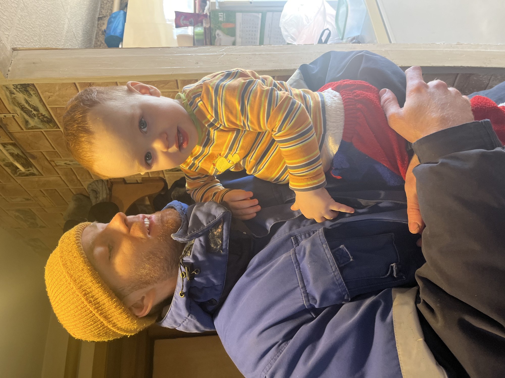 A medium shot captures a man lying on his back with a young child resting on him. The man is fair-skinned and bearded, wearing a yellow beanie, a blue jacket, and smiling. The child, with fair skin and reddish-brown hair, is wearing a striped orange and yellow shirt and red pants. The child is looking towards the camera. The background features a tiled floor. This image was taken in a domestic environment, likely a residence, in the settlement of Сосново, Russia.