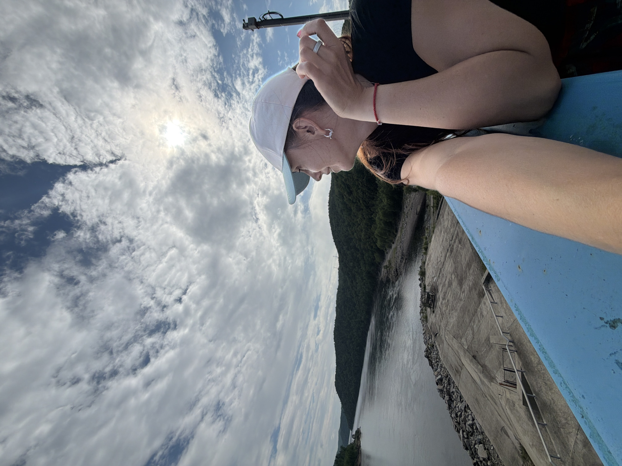 The image shows a woman wearing a white and blue baseball cap and a black t-shirt, looking out at a wide river and densely forested hills under a cloudy sky with the sun peeking through. She is leaning on a blue railing. The foreground shows a concrete structure with stairs leading down towards the river. The location is identified as the Talakan Hydroelectric Power Station in Talakan, Russia.