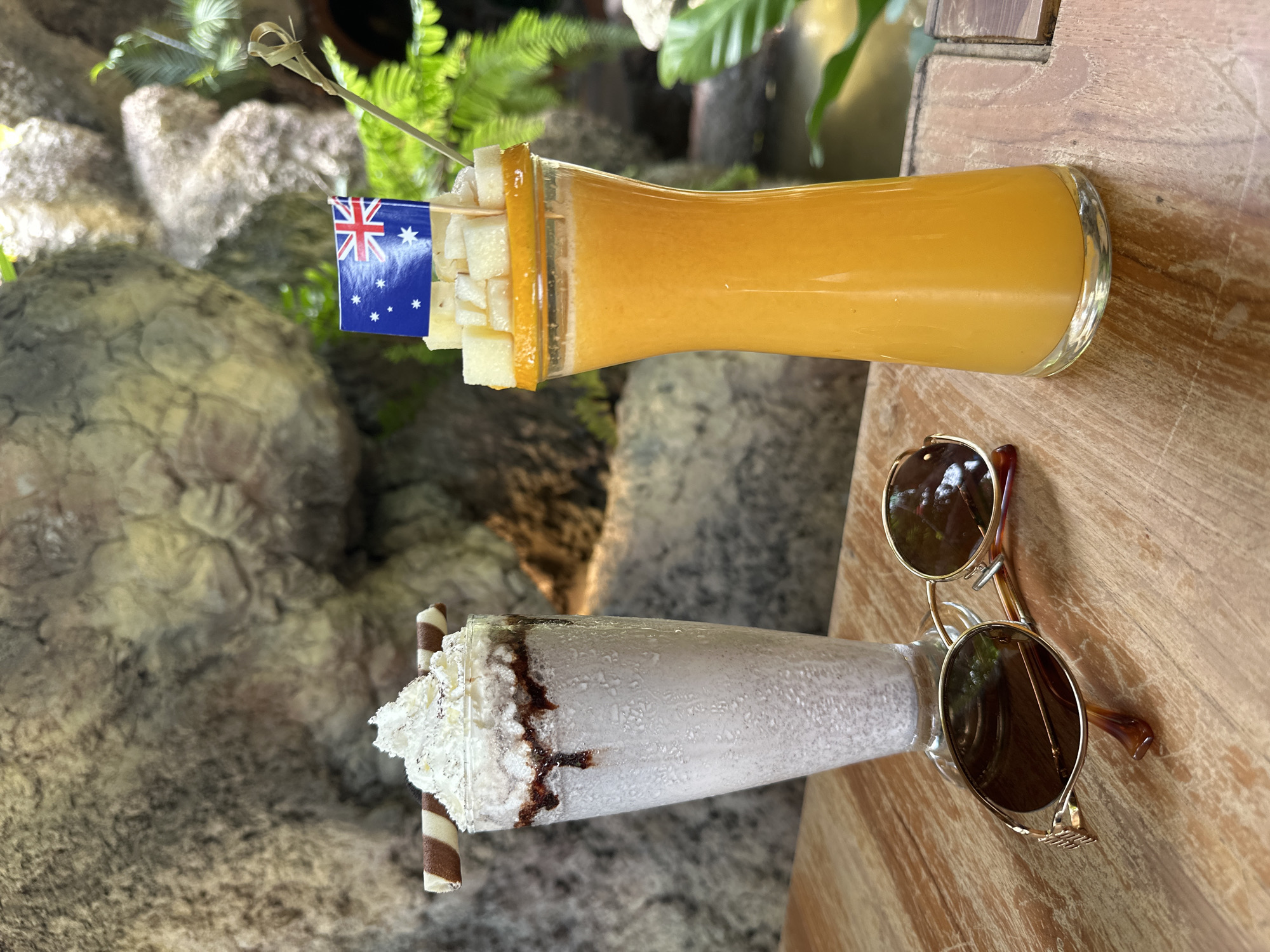Two drinks are on a wooden table in an outdoor setting, likely in Ko Samui District, Thailand, as suggested by the tropical foliage and rock formations in the background, resembling a resort or restaurant patio.
The drink on the left is a tall, frosted glass filled with a pale, greyish-white beverage, possibly a milkshake or smoothie. It is topped with a generous swirl of whipped cream and drizzled with a dark chocolate sauce. A striped chocolate wafer stick is placed horizontally on top of the whipped cream.
To the right, another tall glass contains a vibrant orange-colored drink, likely a juice or smoothie. The rim of this glass is decorated with thin orange slices. Cubed pieces of fruit, possibly pineapple, are arranged on top of the orange slices. A small Australian flag on a toothpick is inserted into the fruit, and a bamboo skewer is visible behind it.
In front of the drinks, a pair of gold-rimmed sunglasses with dark lenses rests on the table. The sunglasses have tortoise-shell colored arms. The table itself is made of light-colored wood with a visible grain. The background features a textured, rocky wall with patches of lush green foliage, suggesting a natural or landscaped environment. A subtle reflection of green leaves is visible in the sunglasses.