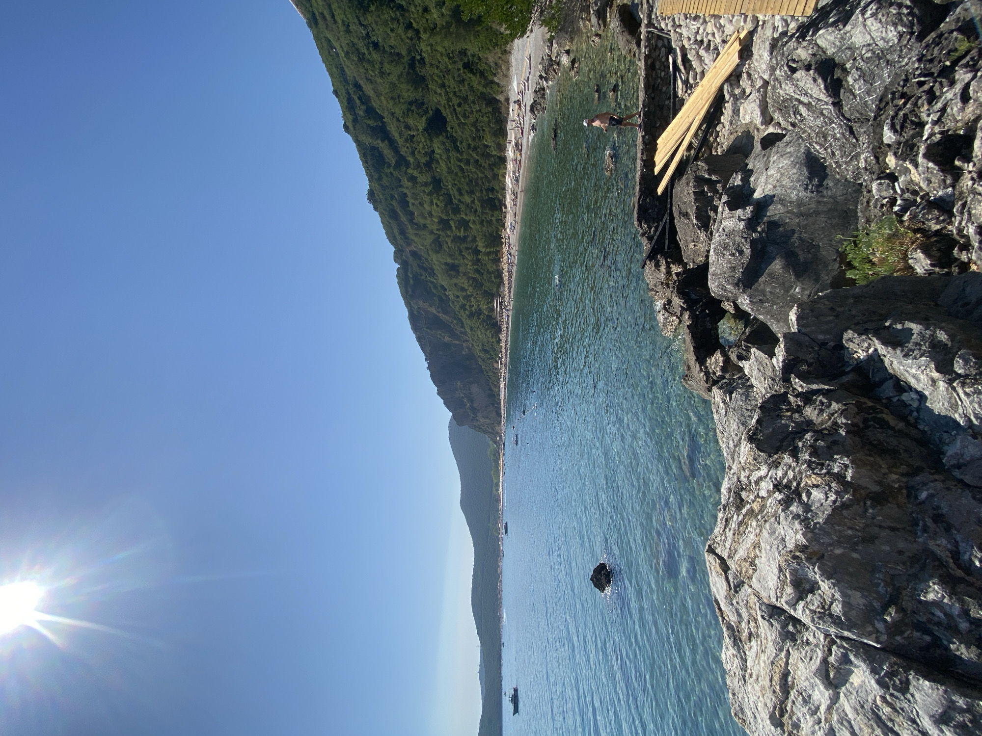 A wide-angle, eye-level, outdoor, daylight, sunny, horizontal, medium shot captures a picturesque view of a rocky coastline along a calm, clear turquoise sea. The sun is high in the bright blue sky, casting a lens flare in the upper left corner. In the foreground, large grey and textured rocks dominate the lower portion of the frame. To the right, a wooden walkway or structure is under construction, with several planks of wood stacked neatly. A person, a male wearing a white hat and dark shorts, stands on a low stone wall, looking out towards the sea.
The middle ground features the expanse of the sea, with visible underwater rocks and pebbles. A few small boats are scattered across the water, and a dark rock protrudes from the sea near the left side.
The background reveals a densely forested, green hillside that slopes down towards a long, pebble beach. This beach is lined with numerous white beach umbrellas and lounge chairs, indicating a popular recreational area. The coastline curves into the distance, with more hazy green hills forming the backdrop under the clear blue sky. The location is identified as being near Prijevor, Montenegro.