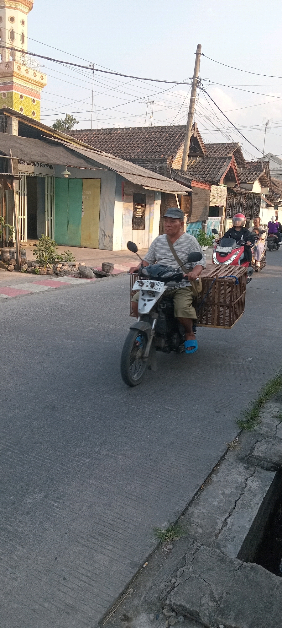 A man wearing a striped t-shirt, olive-colored pants, blue flip-flops, and a dark grey cap is riding a motorcycle with a wooden cage attached to the back. He is in motion on a paved road, likely in the city of Babat, Indonesia, as indicated by the presence of a minaret-style building and buildings with traditional Indonesian architecture. The man is looking forward, his expression neutral. Behind him, other motorcycles are also on the road, with people riding them. The environment appears to be a street scene with a sidewalk, some vegetation, and utility poles with numerous wires. The lighting suggests it is either early morning or late afternoon.