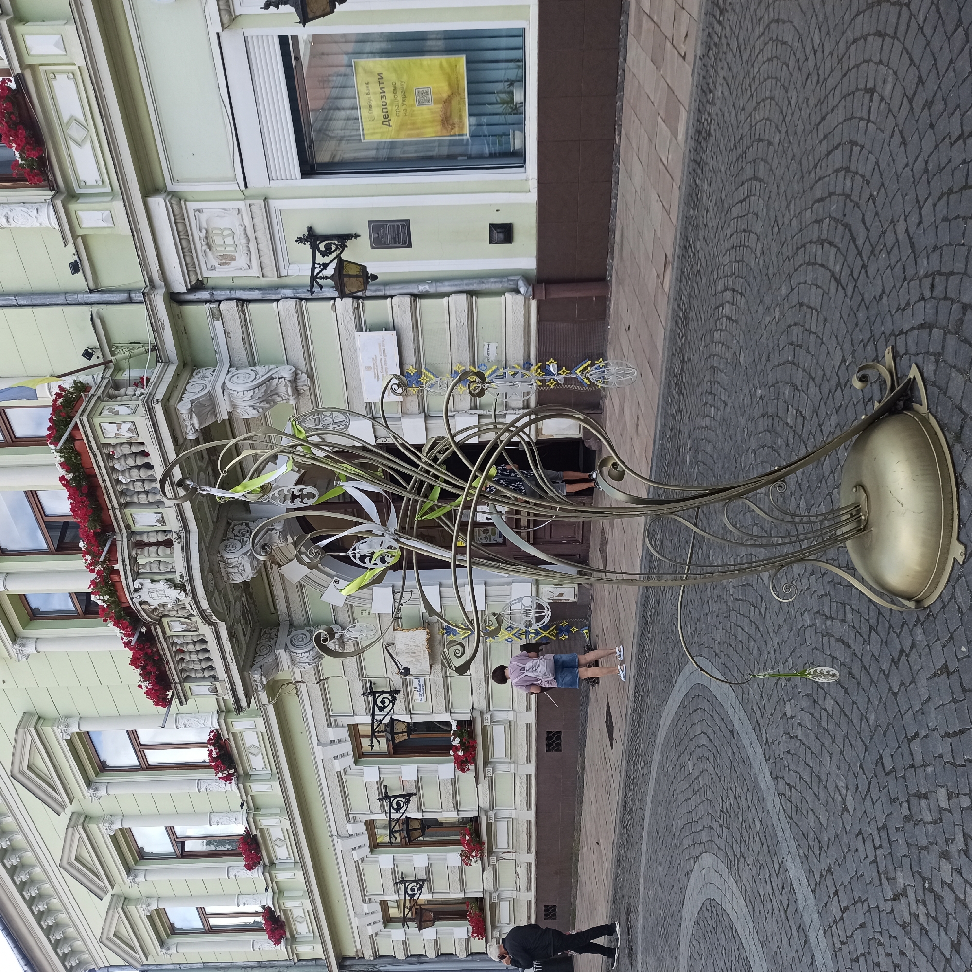 The image depicts an outdoor scene on a cobblestone street in Chernivtsi, Ukraine. In the foreground, a large, ornate golden sculpture with flowing, organic shapes is prominently displayed. This sculpture is adorned with delicate white and green ribbons and small, globe-like ornaments. The background features a light green building with a classical architectural style, characterized by columns, pediments above windows, and a decorative balcony. Flower boxes filled with red and white flowers are visible on the balcony and window ledges.
Two individuals are present. To the left, a person wearing a dark jacket and pants is walking away from the camera, holding a phone to their ear. To the right, a person with short dark hair, wearing a purple t-shirt, denim shorts, and a backpack, stands facing the building and holding what appears to be a stick or a staff.
On the building's facade, several signs are visible. One sign in a shop window displays the logo of "Globus Bank" and text in Ukrainian, and another sign on the exterior of the building has text in Ukrainian as well. Street lamps with a black, ornate design are mounted on the building. The street itself is paved with dark gray cobblestones, arranged in a circular pattern, with a lighter gray paved area in the distance. The overall atmosphere suggests a public space, possibly a square or a pedestrian area in the city.