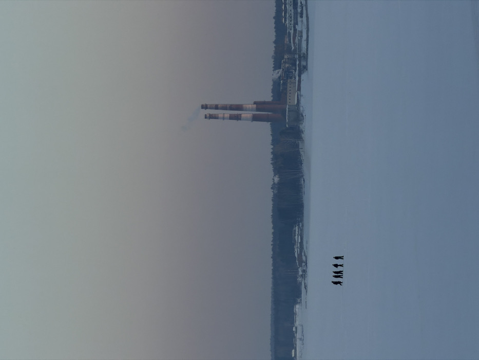 The image displays a panoramic winter landscape on the Votkinsk Reservoir in Chaikovsky, Russia. Five individuals are visible in the mid-ground, presented as dark silhouettes, walking in a staggered line from left to right across the flat, snow-covered surface of the frozen reservoir. In the distant background, beyond a dark line of dense forest and scattered buildings on the far shore, the Chaikovsky Thermal Power Plant is identifiable by two tall, slender industrial chimneys. These chimneys are vertically striped with alternating light and dark bands, and faint vapor emanates from the top of the left chimney. The sky above is uniformly overcast, exhibiting a subtle gradient from light blue-grey at the zenith to a darker purplish-grey closer to the horizon. The scene is illuminated by diffused, cool light, characteristic of a low-light winter environment.