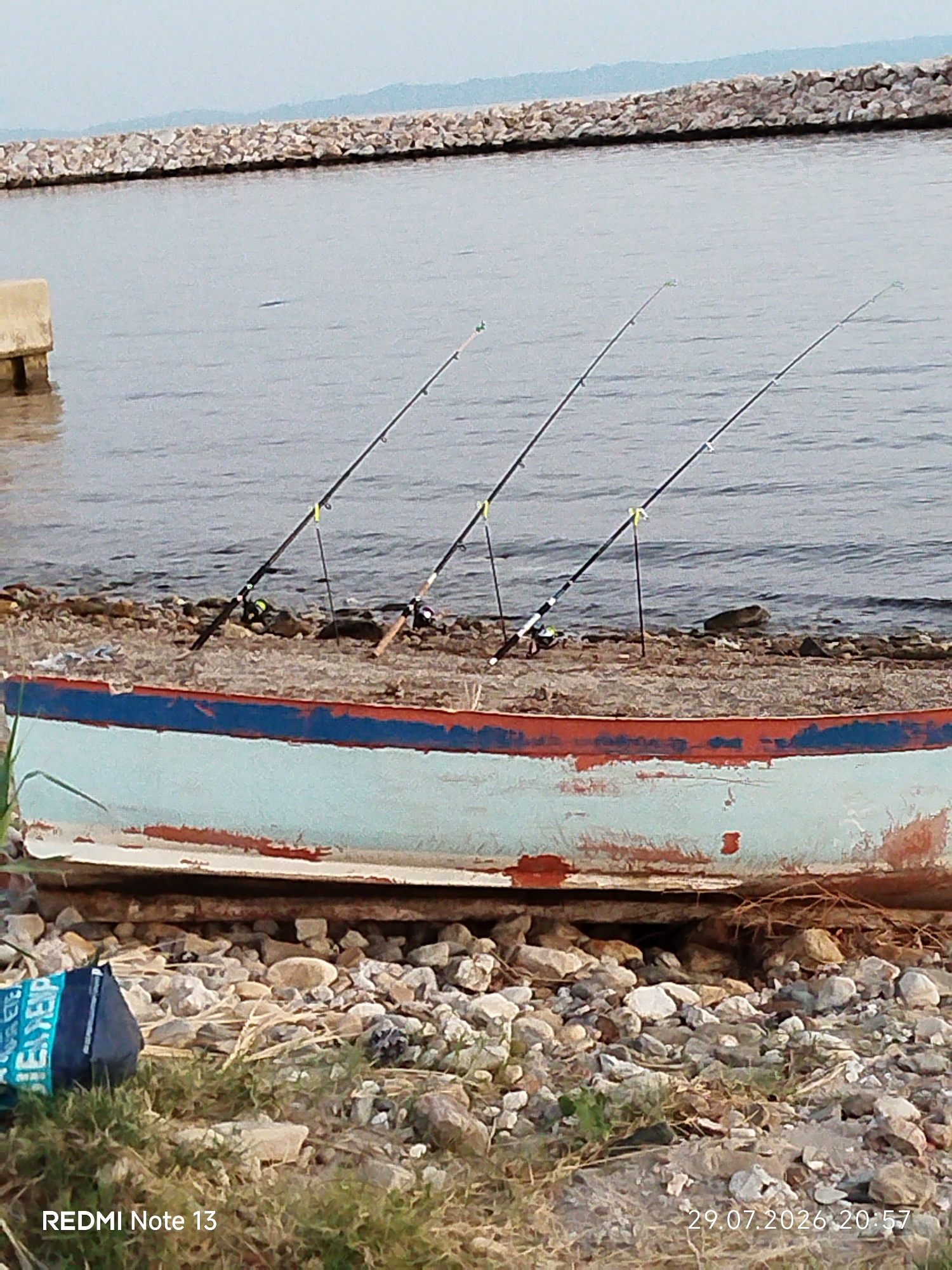 The image depicts a scenic coastal view in the Sithonia Municipal Unit, Greece. In the foreground, an old, weathered boat with chipped blue and red paint rests on a rocky shoreline. Three fishing rods are planted upright in the pebbles, their lines extending towards the calm, grey water. The sea stretches to the horizon, meeting a hazy sky. In the distance, a man-made stone breakwater juts out into the water, with a concrete structure visible on the left side of the frame, possibly a pier or dock. The overall atmosphere is tranquil and serene, suggesting a peaceful day of fishing.