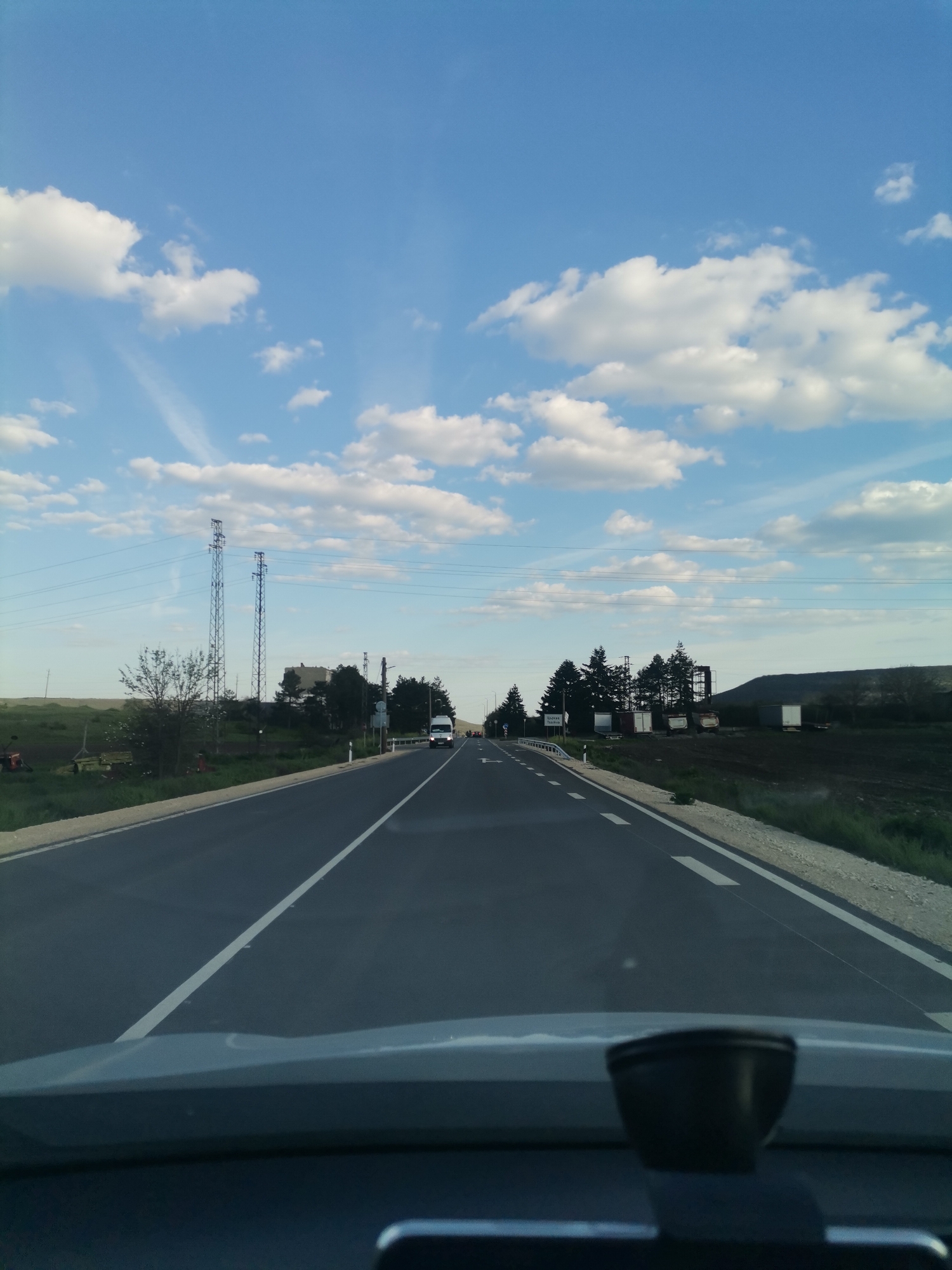 The media file presents a view from inside a vehicle, looking forward down an asphalt road on a clear day with scattered white cumulus clouds against a bright blue sky. The time of day appears to be late afternoon or early evening due to the soft lighting.
The road features a solid white line dividing the lanes and broken white lines along the right shoulder. To the left, the landscape consists of green fields, some patches of dark, cultivated earth, and two tall metal electricity pylons supporting power lines that traverse the scene. Further left, trees and what might be agricultural or industrial structures are visible, with a small red tractor partially obscured in the far distance.
On the right side of the road, there is a gravel shoulder, followed by a line of evergreen trees. Beyond the trees, several white container-like structures are present, backed by a dark, elevated landform, possibly a hill or a spoil heap. An oncoming white van with its headlights on is visible in the distance.
The immediate foreground shows the dashboard of the car, with a dark, rounded object, likely a dashcam or sensor, mounted on it. A small, white road sign on the right, partially hidden by trees, bears the Cyrillic text "C. ЦАРКВА," indicating the location as Tsarkva, Bulgaria. The scene depicts a drive through a rural or semi-rural area. No individuals are explicitly visible, though the white van suggests other road users.