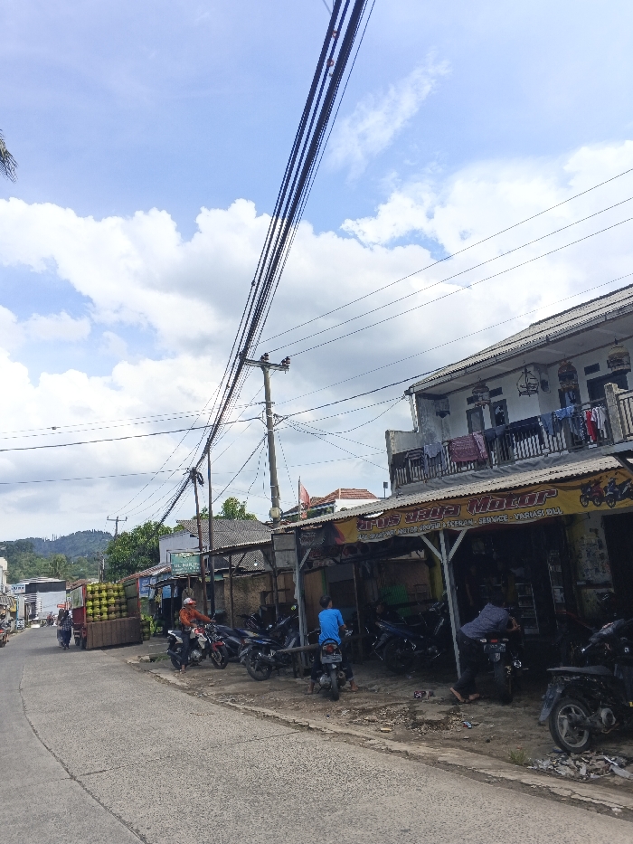 The image depicts a daytime street scene in the city of Cordon, Philippines. The sky is bright blue with scattered white cumulus clouds. A paved road, appearing somewhat worn, curves gently to the left in the foreground.
On the right side of the road, a two-story building dominates the midground. The ground floor functions as a motorcycle repair and variations shop named "AROS JAYA Motor," identified by a prominent yellow banner with black lettering. Multiple motorcycles are parked both inside the open-front shop and on the unpaved area in front. Two male individuals are present in this area: one, in a blue t-shirt and dark pants, is seated on a motorcycle facing away from the camera; another, in a patterned shirt, is barefoot and bending over a motorcycle, appearing to perform maintenance. The upper floor of the building has a balcony from which laundry is hanging on a line. Several ornate, dark-colored bird cages are suspended from the building's facade at the second-story level.
Numerous wooden utility poles with a dense network of overhead electrical and communication wires extend across the upper portion of the scene, crossing above the street and buildings.
On the left side of the road, further down, a large stack of green, spherical produce, consistent with coconuts, is visible on what appears to be a cart or small truck. Nearby, a male individual wearing an orange shirt and a white helmet is seated on a motorcycle, facing towards the camera.
In the background, behind the structures, green hills or mountains are visible under the horizon line. The overall environment suggests a semi-urban commercial area with active daily life and commerce.