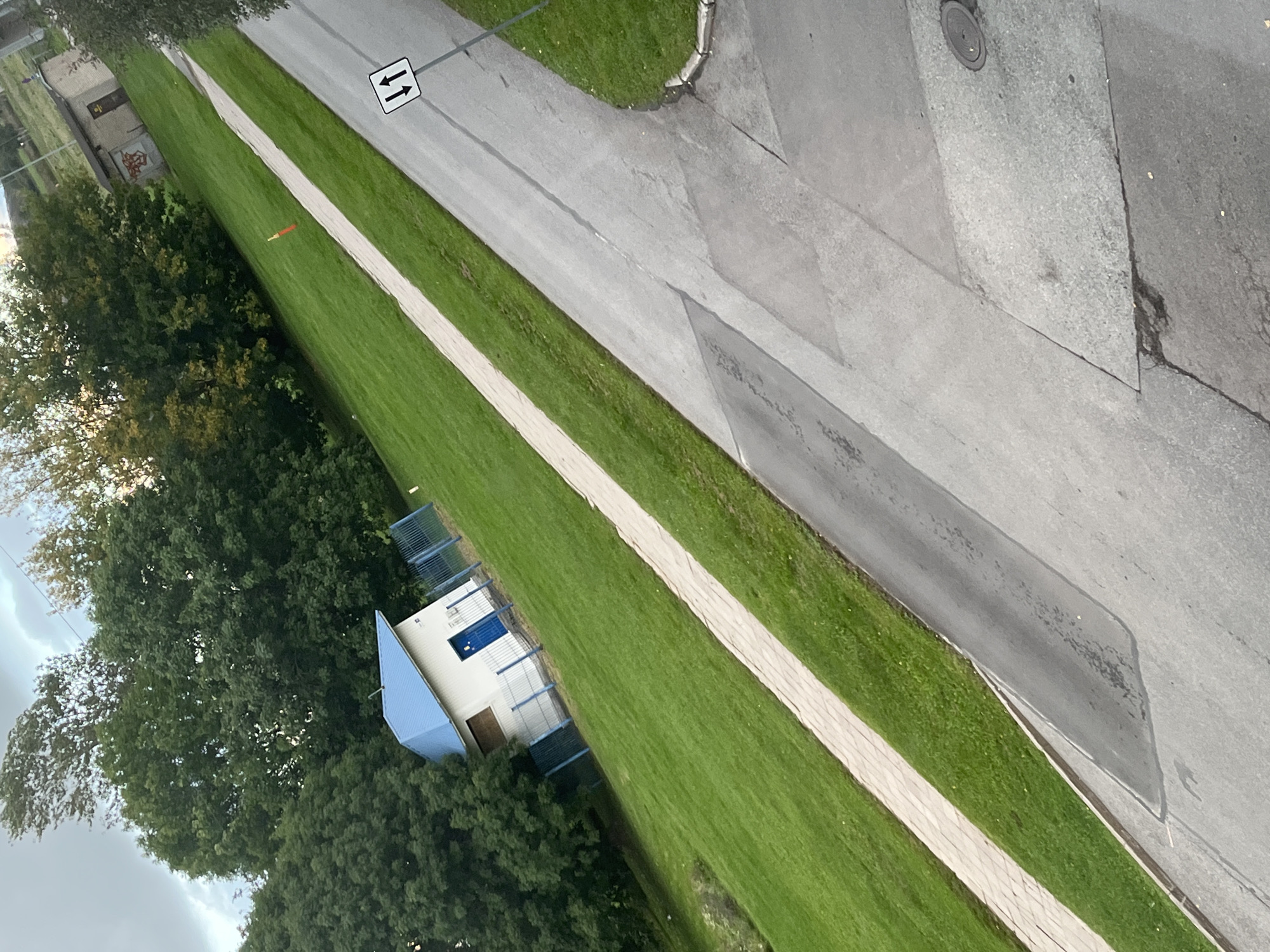 The image shows an outdoor scene with a paved area in the foreground, a grassy slope with a paved walkway, a small white building with a blue roof and door, and trees in the background. There is also a sign with two arrows pointing up and down, indicating two-way traffic. The location is identified as Kohtla-Järve linn, Estonia. The environment appears to be a park or a residential area. The sky is overcast with some clouds. There are no people visible in the image.