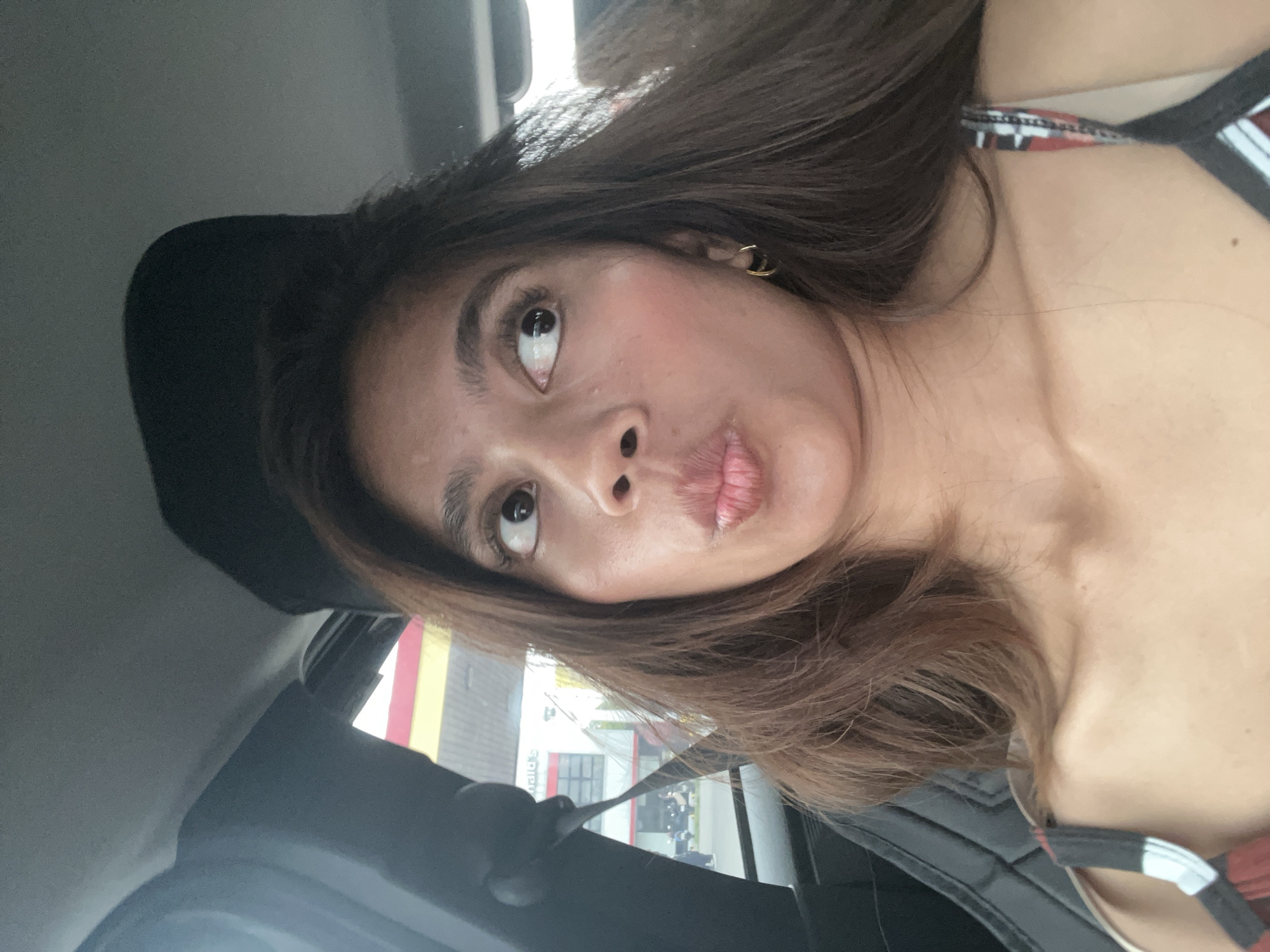 The image shows a close-up selfie of a young woman inside a car. The woman has long, brown hair and is looking to her left with her lips pursed. She is wearing a tank top with a patterned strap. The interior of the car is visible, with a black seat and headrest. Outside the car window, a McDonald's restaurant with its distinctive red and yellow signage is visible. This location appears to be a McDonald's branch in Panabo City, Philippines, given the context provided. The lighting suggests it is daytime.
