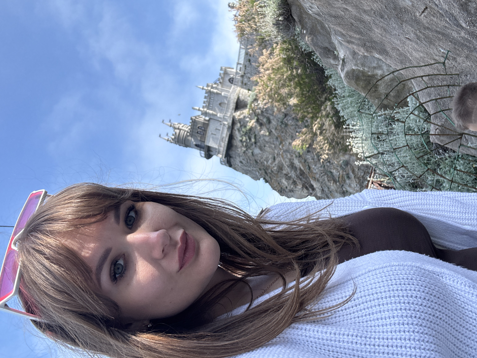 A woman with brown hair and sunglasses is posing for a selfie in front of Swallow's Nest castle located in the city of Ливадийский поселковый совет, Ukraine. She is wearing a white sweater. The castle sits on a rocky cliff with vegetation. The sky is blue.