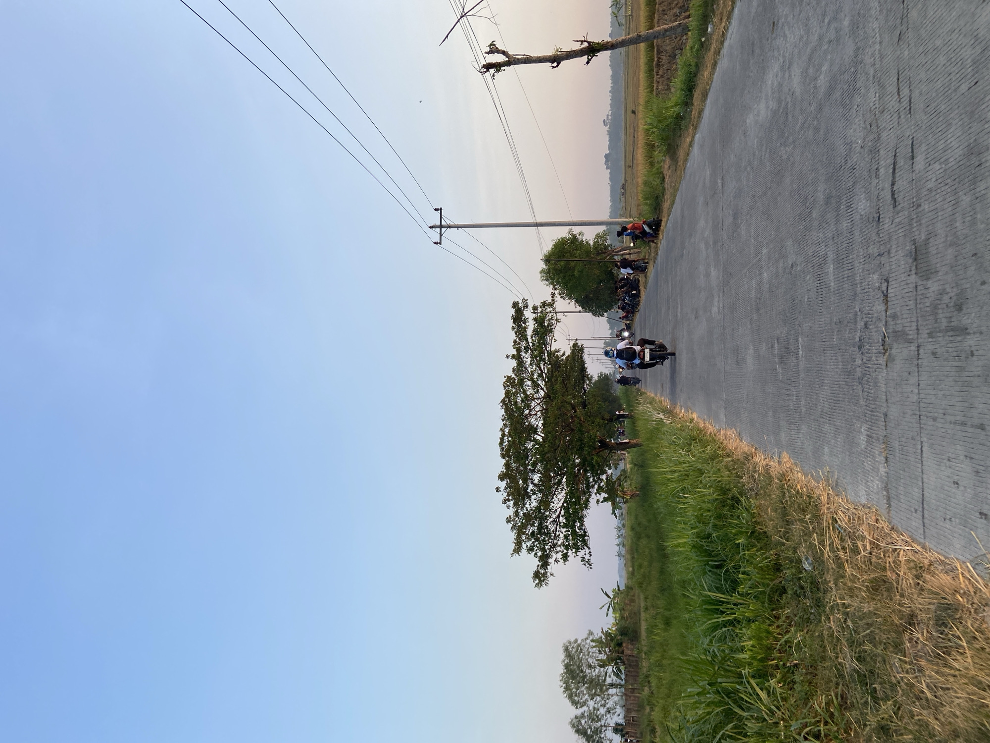The image depicts a concrete road stretching into the distance, flanked by lush green grass and tall reeds on the left, and a harvested rice field on the right. Power lines are strung between utility poles, one of which is prominently featured on the right side of the road. A lone motorcycle rider is heading away from the camera, with a group of people gathered along the side of the road near the motorcycle. In the background, a dense tree stands tall on the left, and a line of trees extends along the horizon. The sky is a clear, pale blue, suggesting either early morning or late afternoon. The scene appears to be set in a rural area, possibly Kropak, Indonesia, given the agricultural landscape and the presence of a local road.
