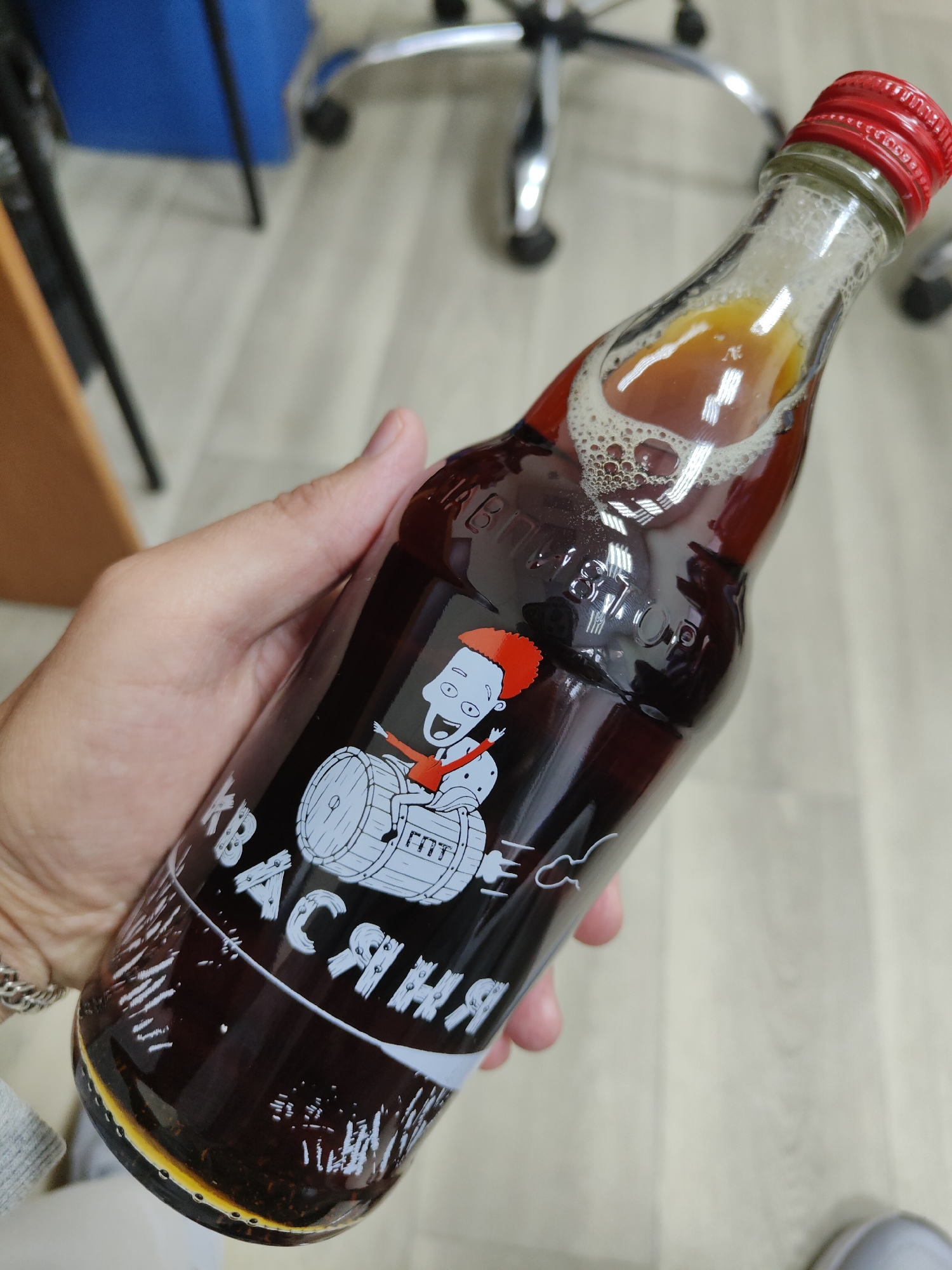 A hand holds a glass bottle of a dark brown beverage with a red cap. The bottle has a cartoon illustration of a smiling person with orange spiky hair sitting on a drum. Text on the bottle is in white and appears to be in Cyrillic script. The background shows an office environment with a light-colored wooden floor and office furniture, including an office chair. The lighting is artificial.