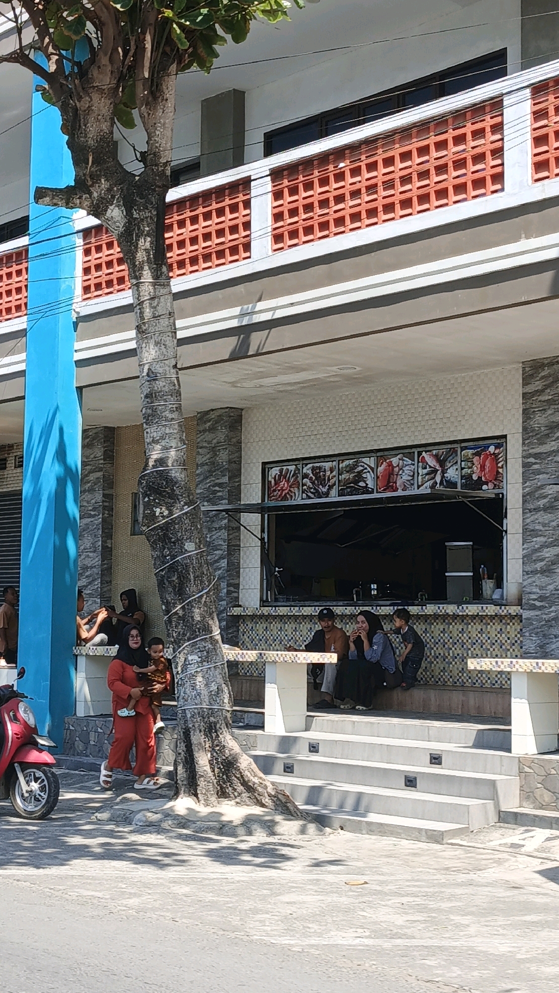 The image captures a street scene in Desa Pagandaran, Indonesia, featuring a building with a prominent blue pillar and a large tree in the foreground. The building has a second-story balcony with orange decorative bricks. In front of the building, several people are seated and standing. A woman in an orange outfit is holding a child. To the right, a group of people are sitting on a bench in front of a window displaying pictures of seafood. A red motorcycle is parked on the left side of the image. The ground in the foreground appears to be asphalt or concrete, with some sandy areas around the tree's base. The scene is illuminated by bright sunlight, casting shadows.