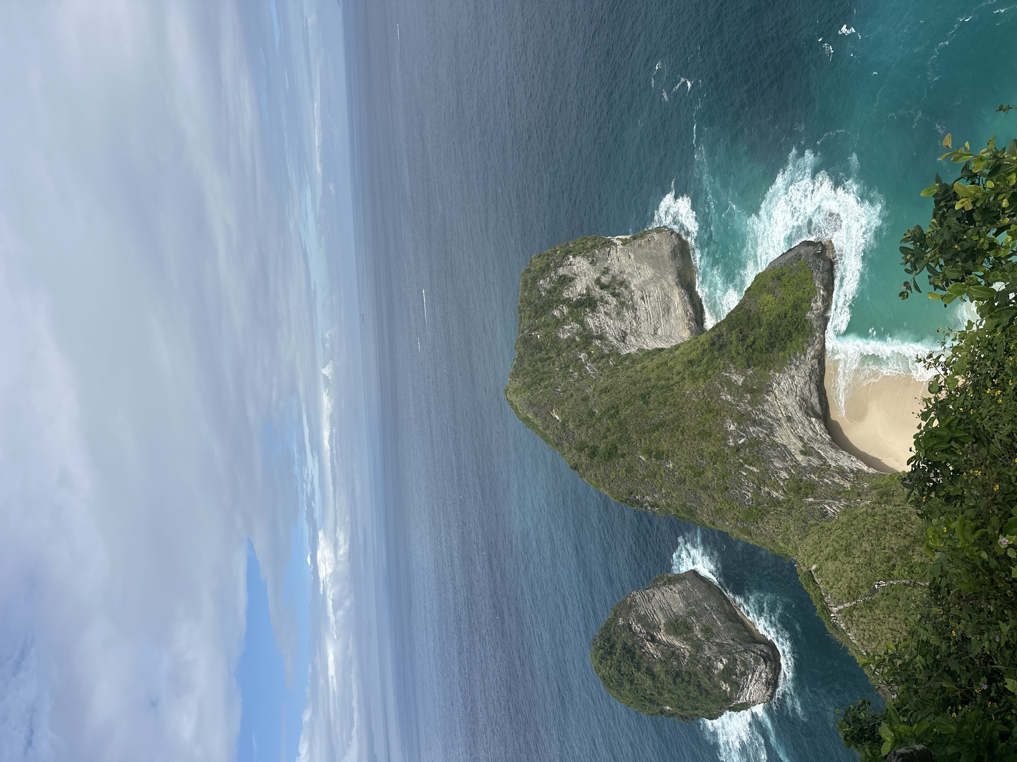 The image shows an aerial view of Kelingking Beach, also known as T-Rex Bay, in Nusa Penida, Indonesia. A headland shaped like a Tyrannosaurus Rex juts out into the turquoise sea, creating a secluded cove with a white sandy beach. Waves crash against the cliffs and shoreline. The ocean stretches out to the horizon, partially obscured by clouds. A smaller rock formation sits to the left of the main headland, also surrounded by waves. Vegetation covers the top of the headland. A narrow trail can be seen winding down to the beach.