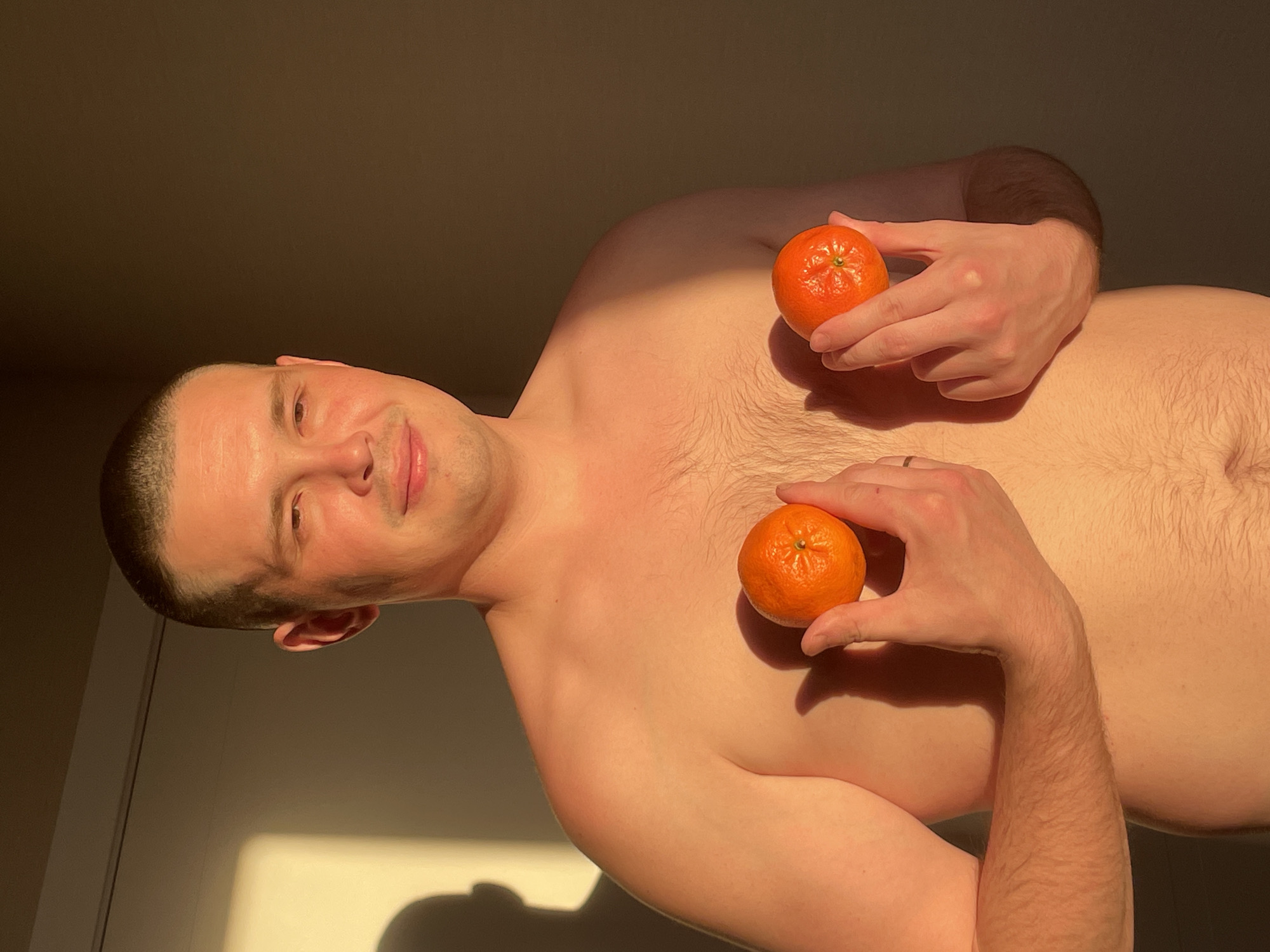 A bare-chested man is holding two tangerines in front of his chest. He has short, dark hair and is smiling. The room is illuminated by a warm light. There is a wall visible in the background. There is no indication of the location being Подшивалово, Russia.