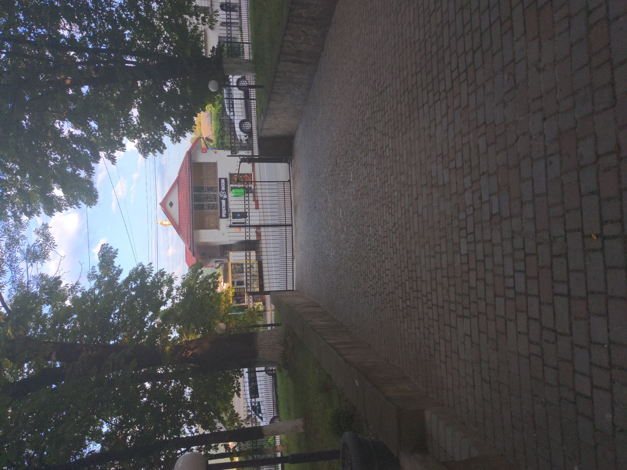 The image depicts a paved walkway leading towards a gated entrance of a building with a red-tiled roof. Two white cars are parked in front of the building, which appears to be a store with signage in Cyrillic. Tall trees with dense foliage frame the scene, and the sky above is partly cloudy. The walkway is constructed of interlocking brick pavers, and a low stone wall runs along the right side of the walkway, separating it from a grassy area. The overall scene suggests an entrance to a commercial establishment in Zalishchyky, Ukraine, possibly a grocery store, indicated by the Cyrillic text on the building's facade.