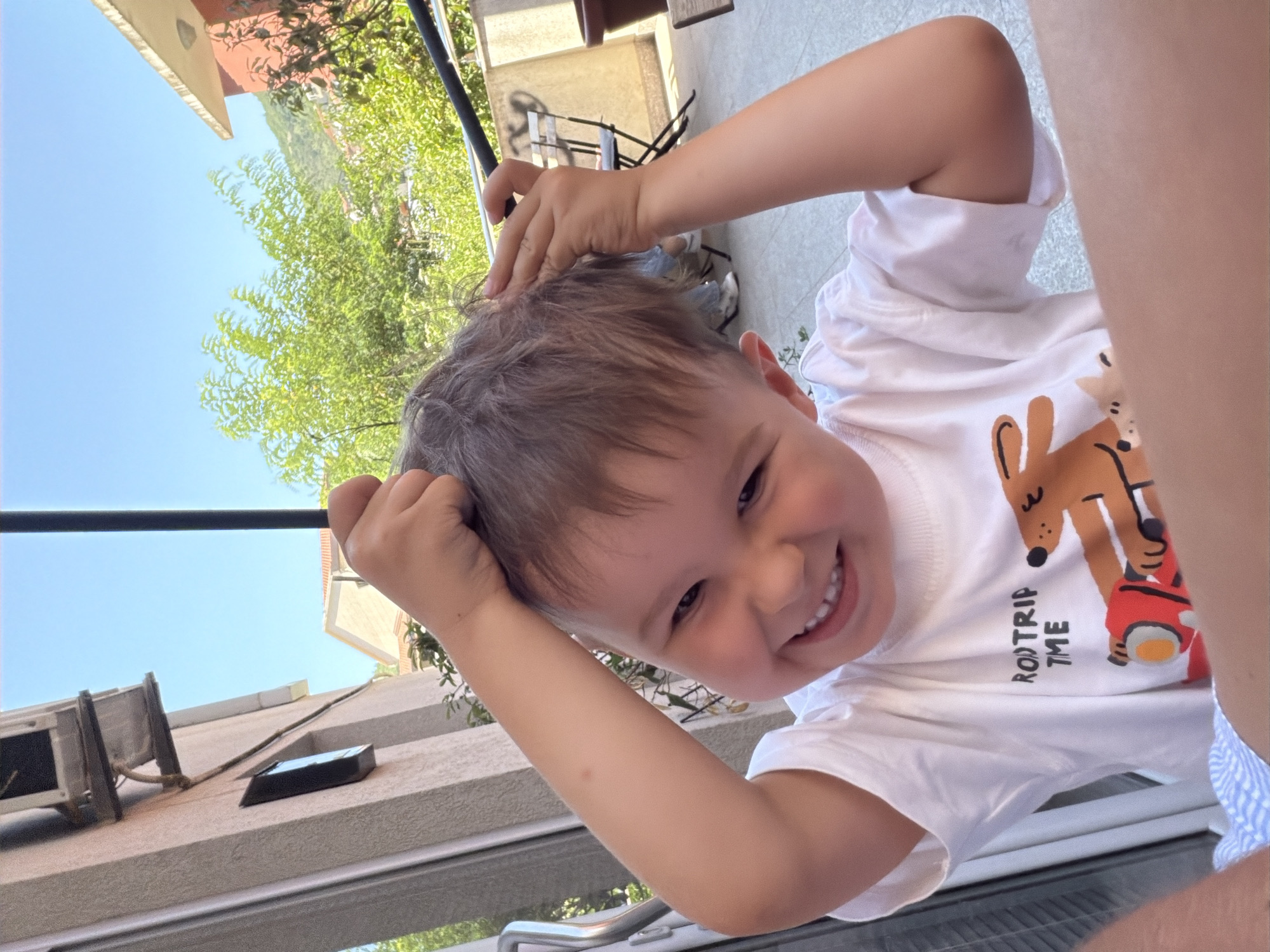 A young boy with light brown hair and a wide smile is pictured from the chest up, standing outdoors. He wears a white t-shirt with a graphic design featuring the text "ROD TRIP TIME" and a cartoon image of a dog with a backpack and a red car. The boy is touching his hair with both hands, lifting strands as if playing with them. He is looking directly at the camera. In the background, there's a building with a light-colored facade and visible windows, suggesting an urban or suburban setting. Lush green trees and a clear blue sky are also visible, indicating a warm, sunny day. The presence of outdoor furniture, such as a chair and table, hints at a patio or balcony. While the exact location is not visible, the architecture and vegetation are consistent with Budva, Montenegro.