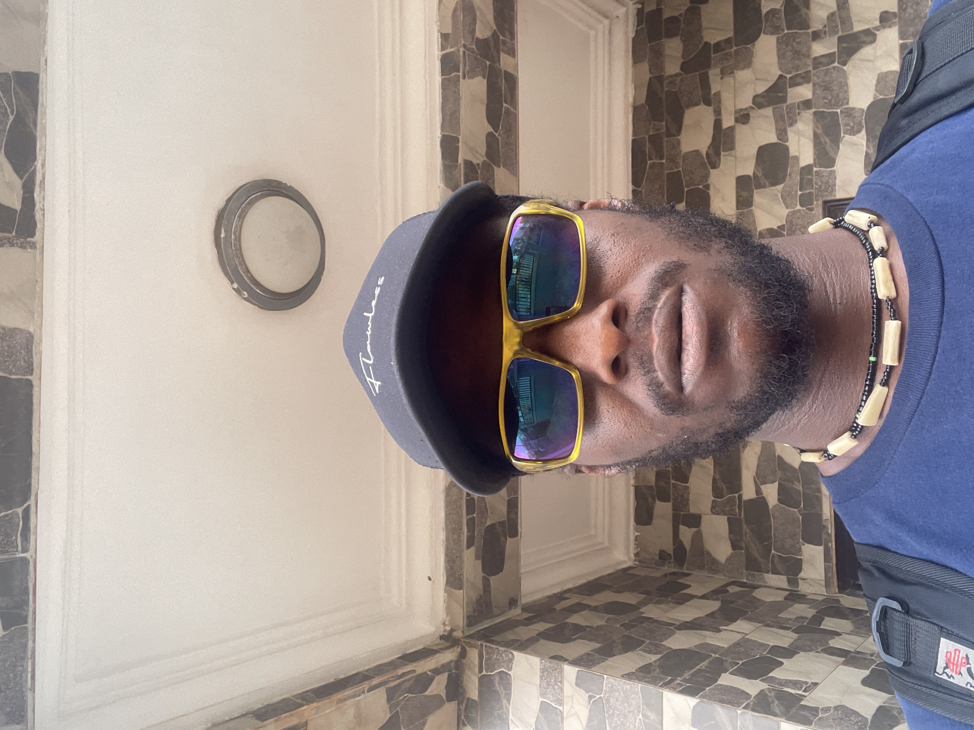 This close-up portrait, oriented sideways in the original, captures a Black man from the shoulders up, likely in a selfie or posed shot. He is wearing a dark blue t-shirt and a black baseball cap with the word "Flawless" embroidered in white script on the front. Large, gold-framed sunglasses with reflective, multi-colored lenses obscure his eyes. He has a well-groomed beard or stubble and his mouth is slightly open, showing a relaxed expression. Around his neck, he wears a multi-strand beaded necklace, featuring small dark beads interspersed with larger, cylindrical off-white beads. A black backpack strap is visible on his right shoulder, featuring a dark buckle and a small brand logo with red elements.
The background suggests an indoor or sheltered outdoor setting, potentially a hallway or porch in Abuja, Nigeria, as per the given context. To the man's left, there is a plain, light-colored wall with a visible decorative trim. On this wall, a round, greyish metallic fixture, possibly a light or doorbell, is mounted. To his right and extending behind him is a patterned tiled wall or column, featuring irregular shapes in shades of grey, brown, and off-white. The lower part of the image reveals patterned tiled flooring with a checkerboard-like arrangement of light and dark grey/brown square tiles. The lighting is bright and even, indicating daytime.